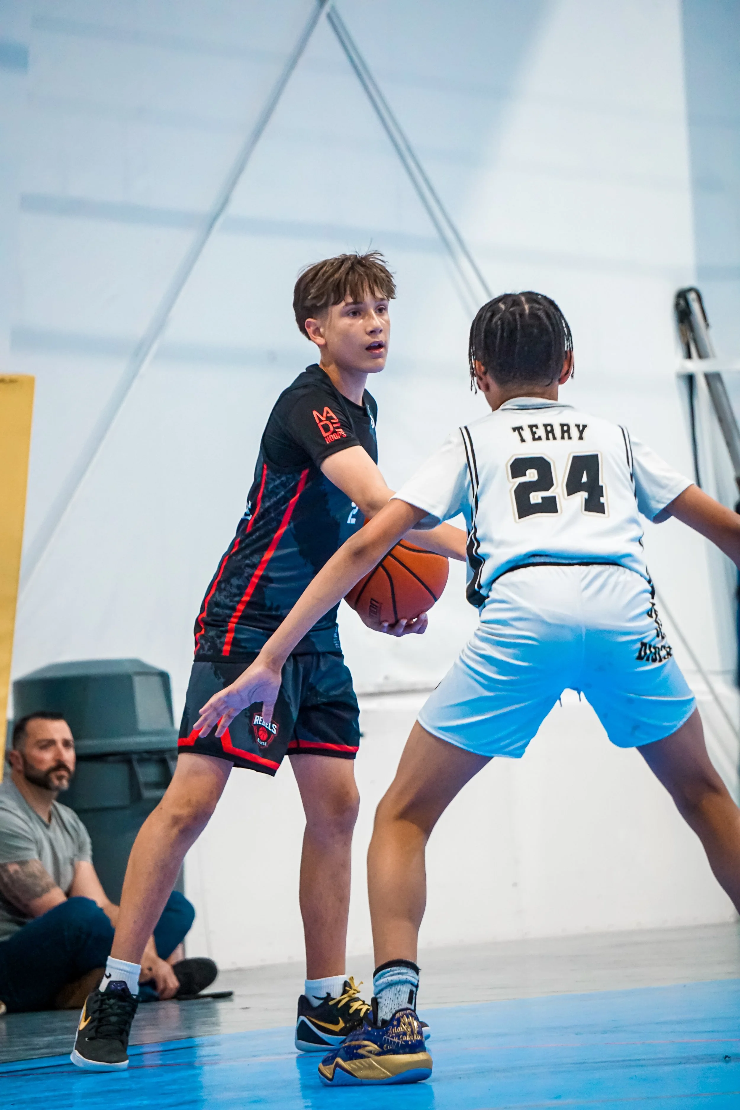 Young boys playing basketball in an indoor gym, with one jumping to shoot or block, and others running and watching.