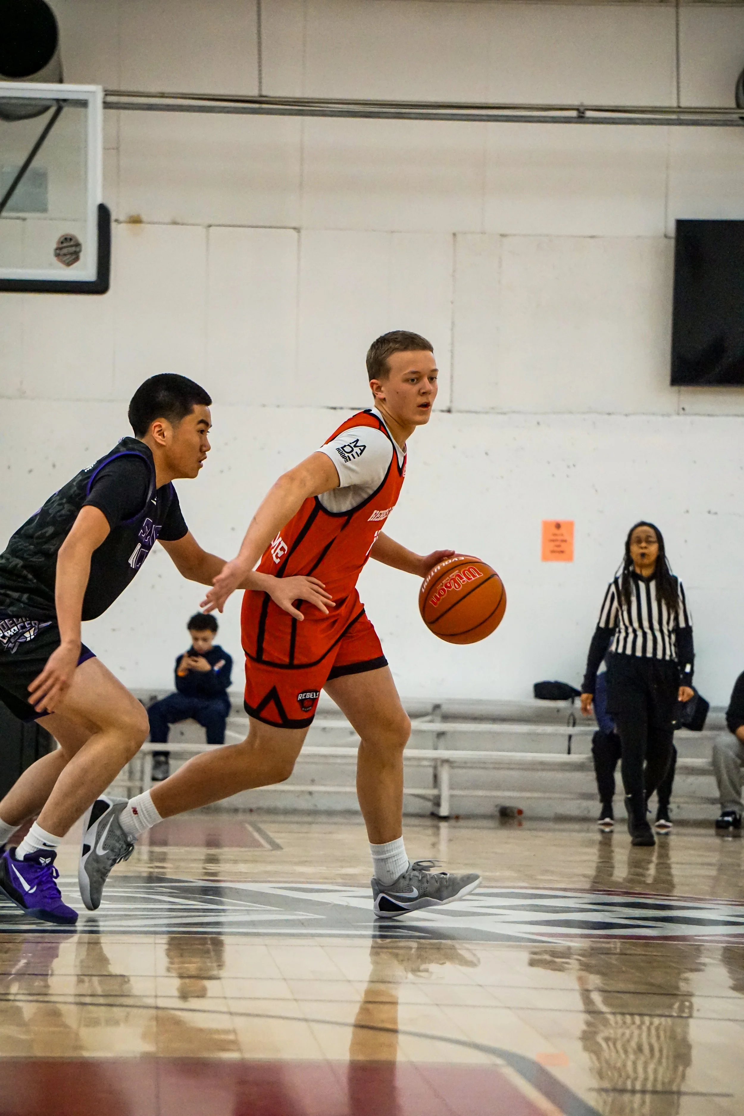 Young male basketball player in white and red uniform holding a basketball on a court, with spectators in the background.