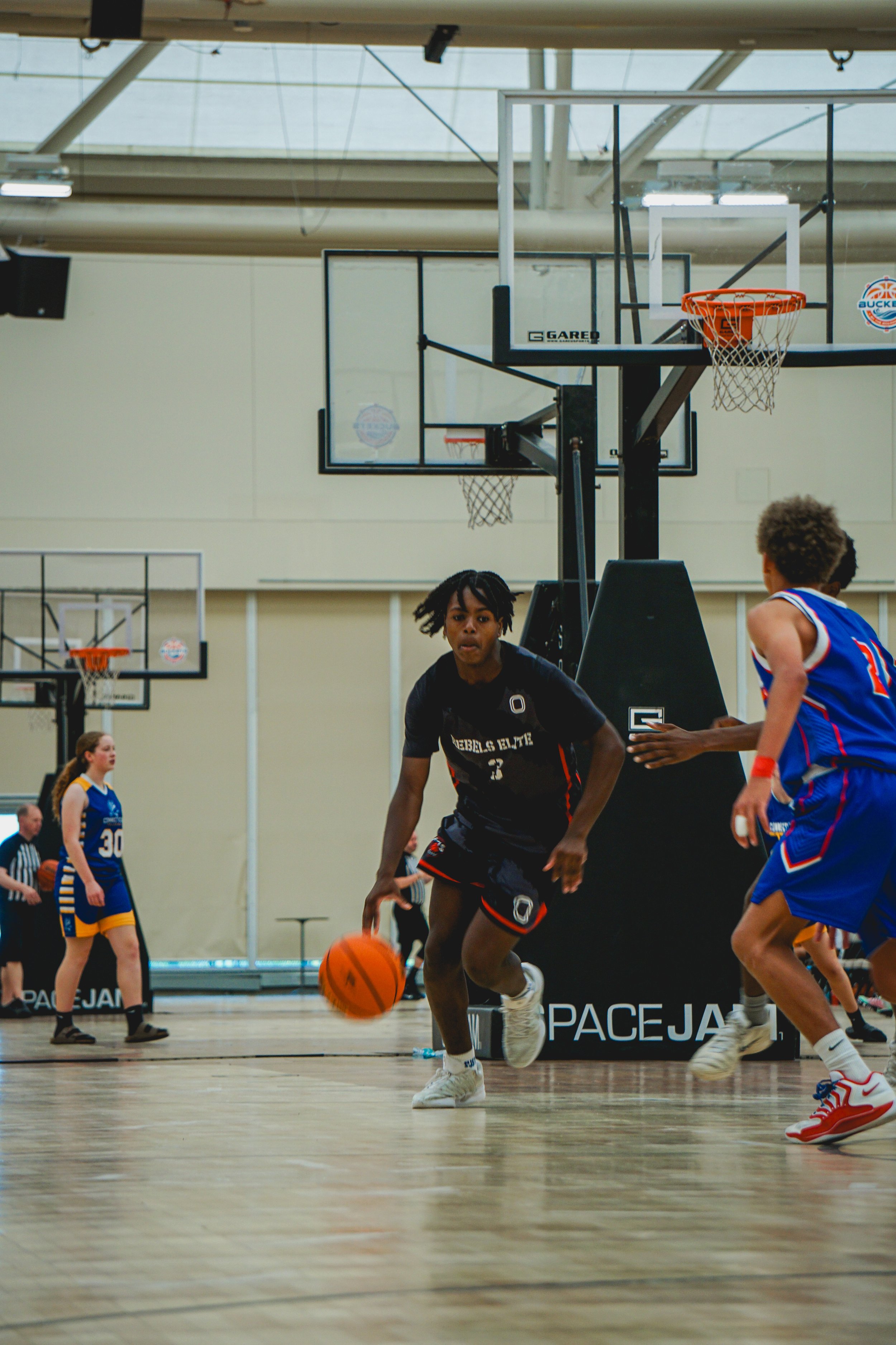 A basketball player wearing a black jersey with red and white accents, number 18, is jumping towards the basketball hoop to make a shot. Other players and spectators are visible in the background inside a gymnasium.