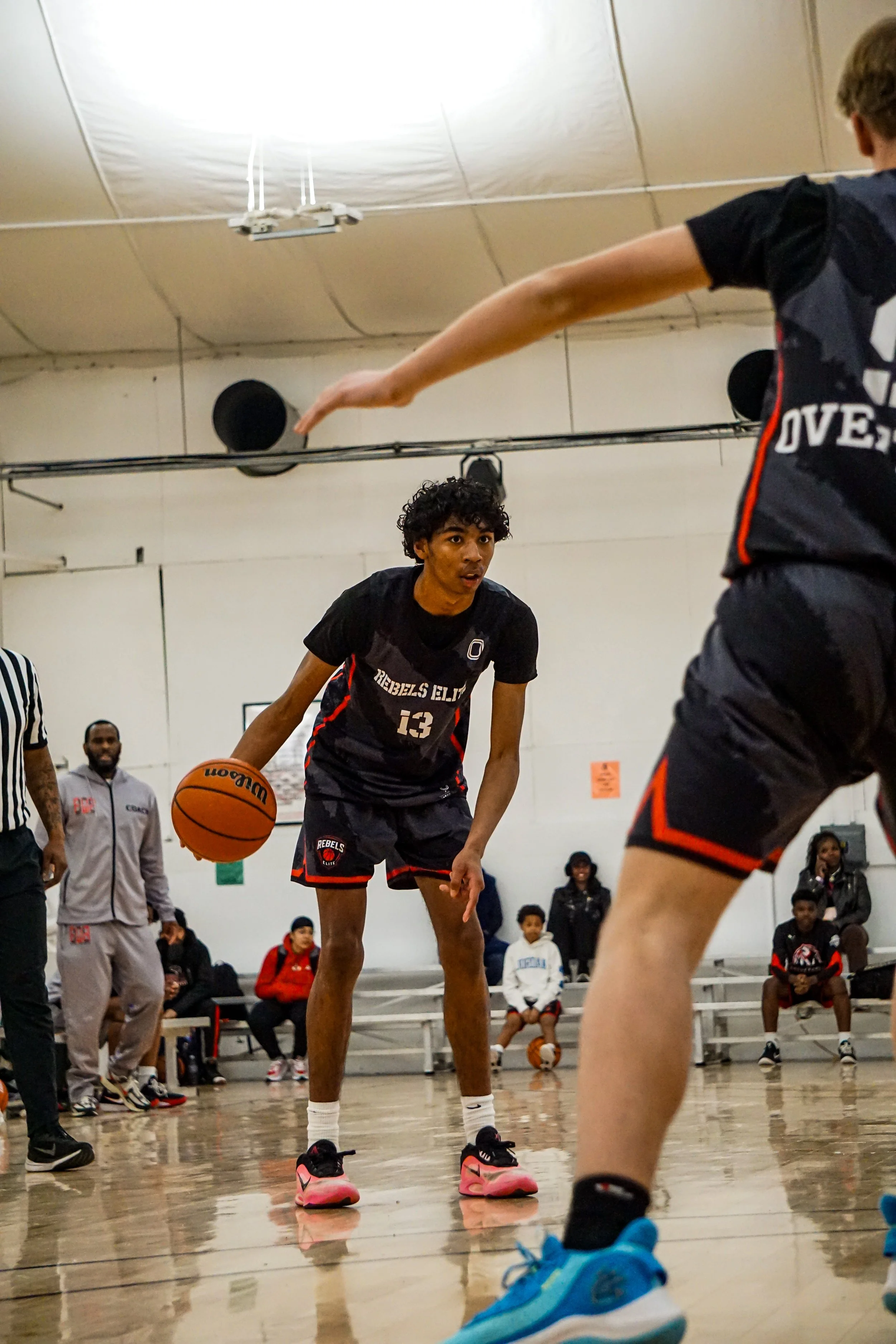 Young basketball player in a white and black uniform dribbling a basketball on an indoor court with teammates sitting on a bench in the background.