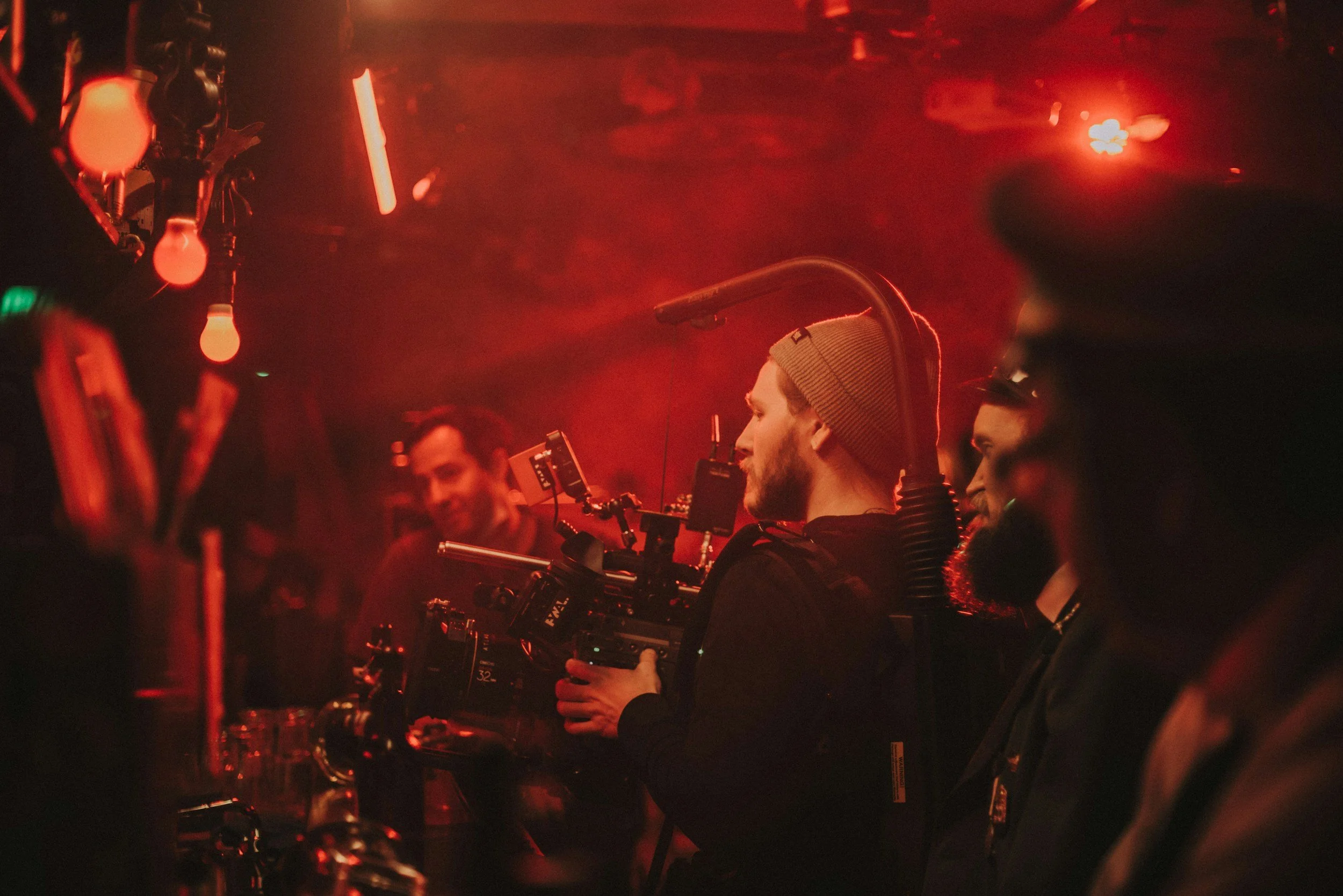A film crew in a dimly lit, red-toned studio. One person is holding a camera rig, wearing a beanie, and is focused on filming. Other crew members are visible in the background, wearing dark clothing and working with equipment.