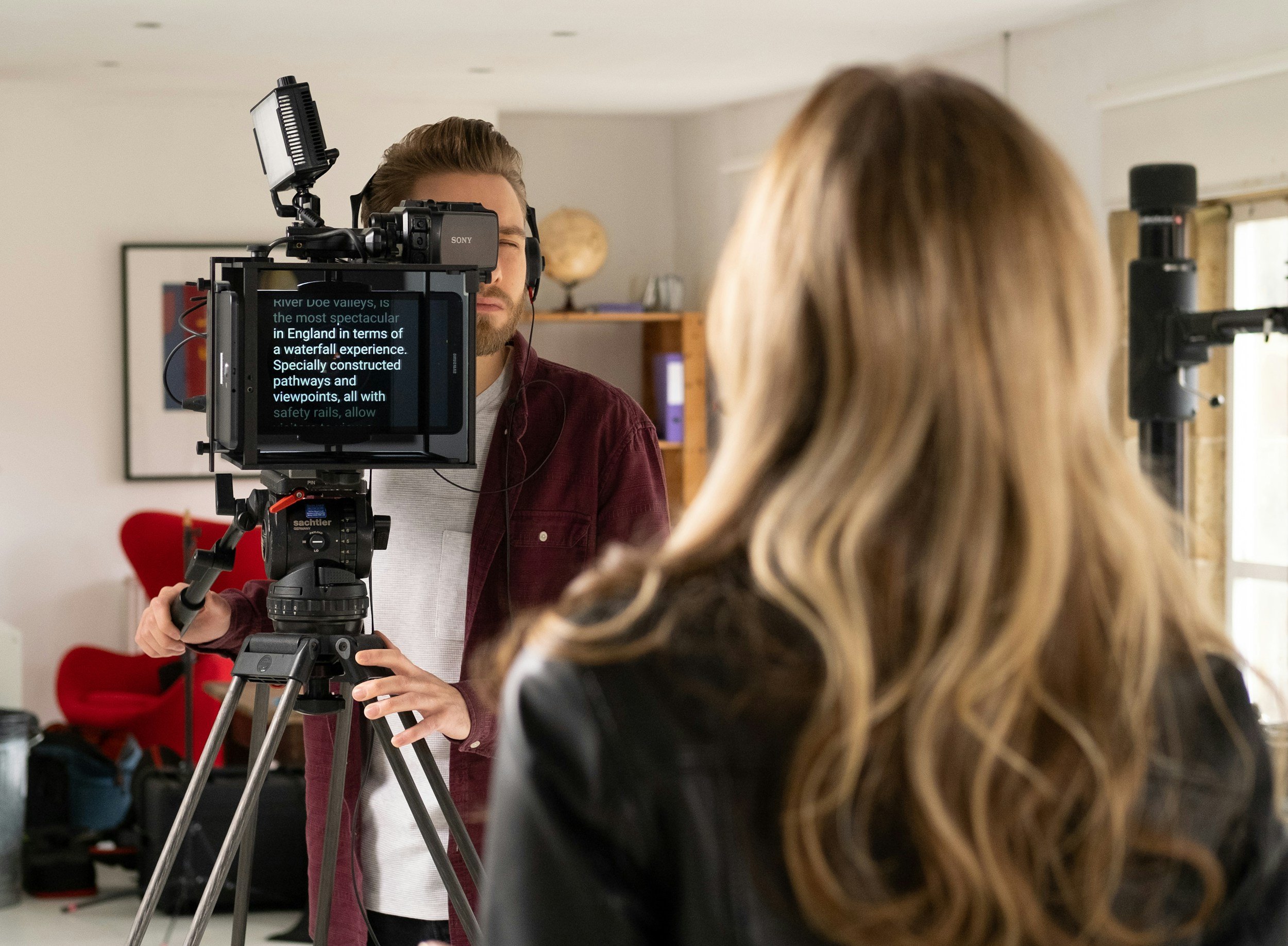 A man operating a professional video camera on a tripod records a woman with long, wavy blonde hair in a room with natural light reading off of a teleprompter.