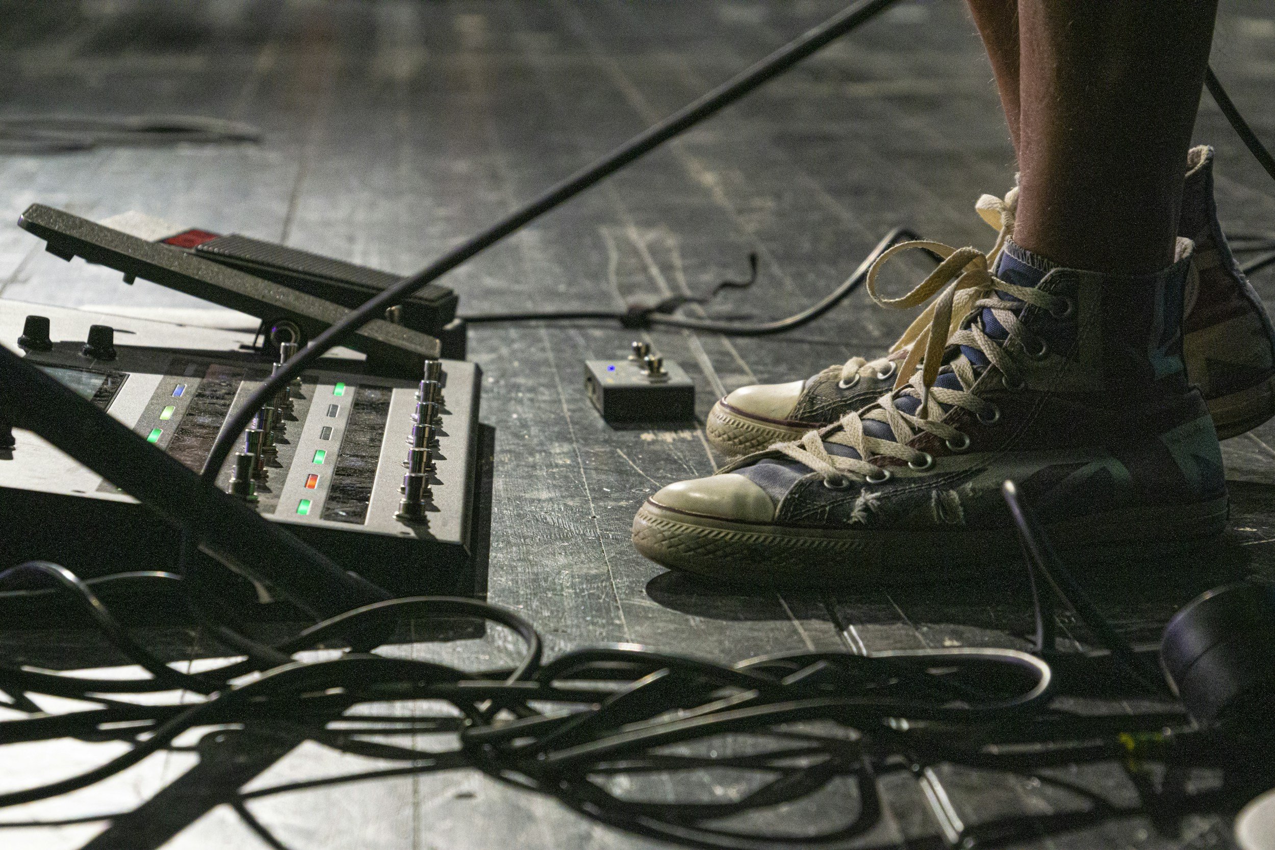 Close-up of person working on a black wooden stage with music equipment, including a pedal, cables, and a multi-effects pedal, surrounding their feet.