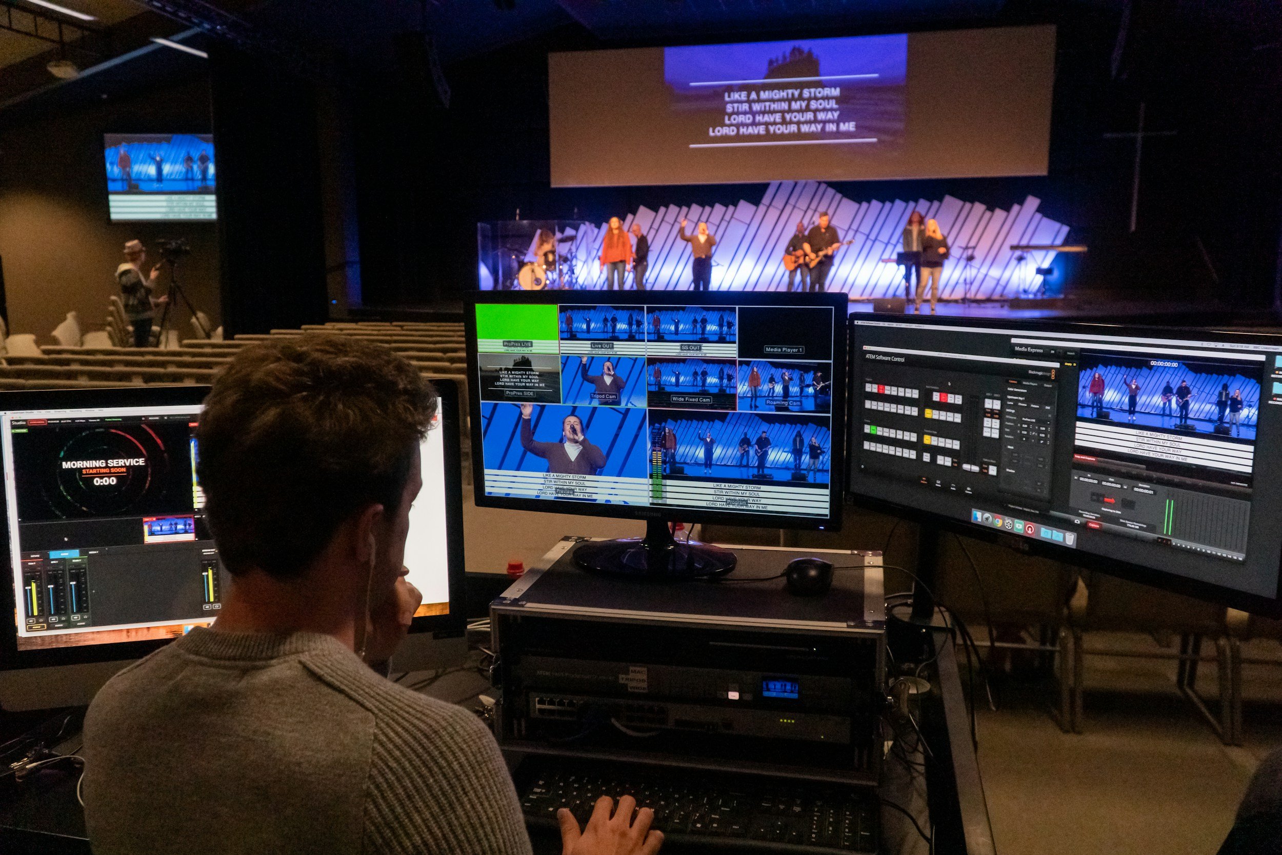A team performs on stage with singers, musicians, and a projected lyric screen in a church or concert hall. In the foreground, a technical crew member operates multiple monitors and a control panel for sound and video editing.