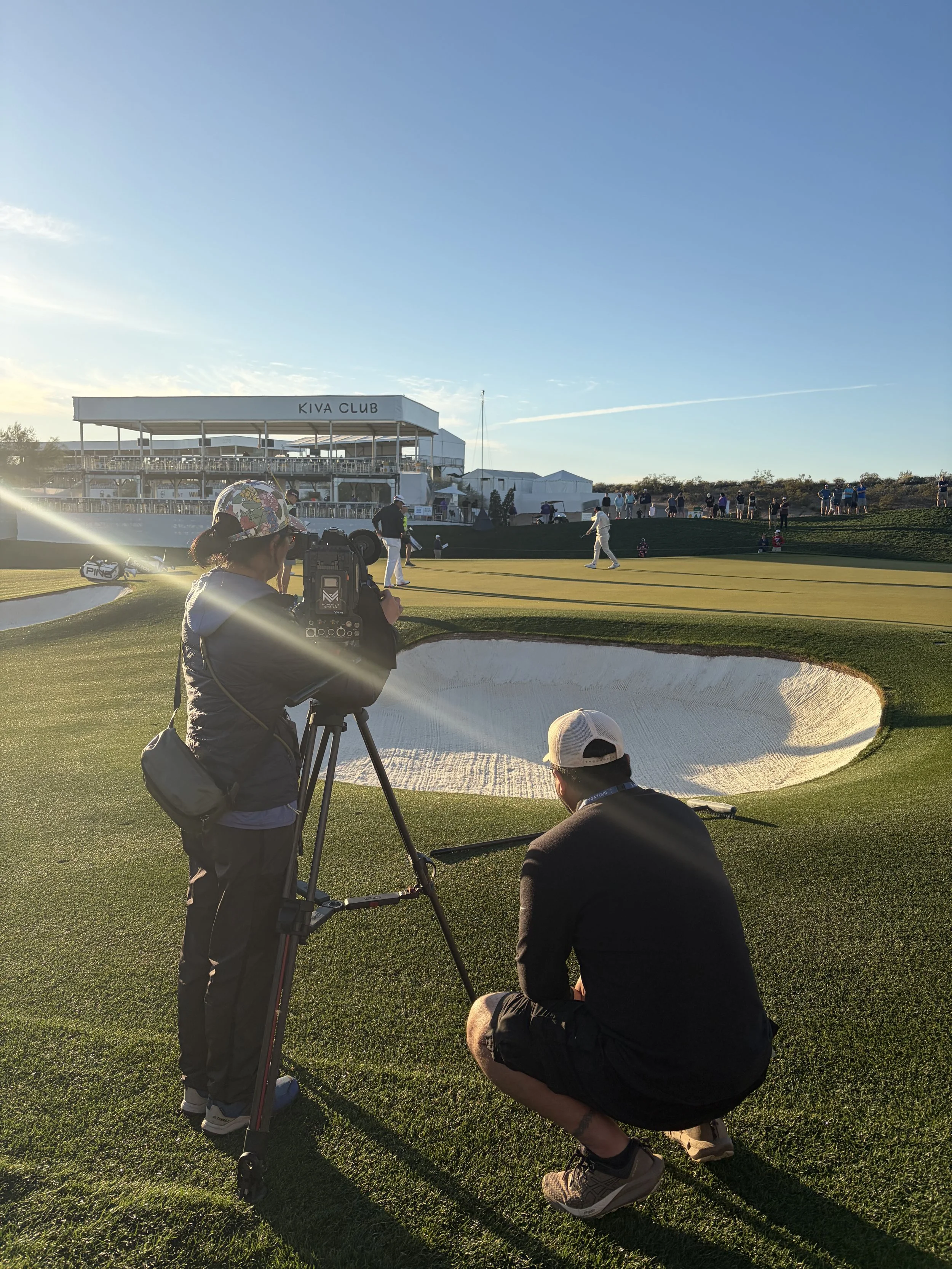 A film crew in a dimly lit, red-toned studio. One person is holding a camera rig, wearing a beanie, and is focused on filming. Other crew members are visible in the background, wearing dark clothing and working with equipment.