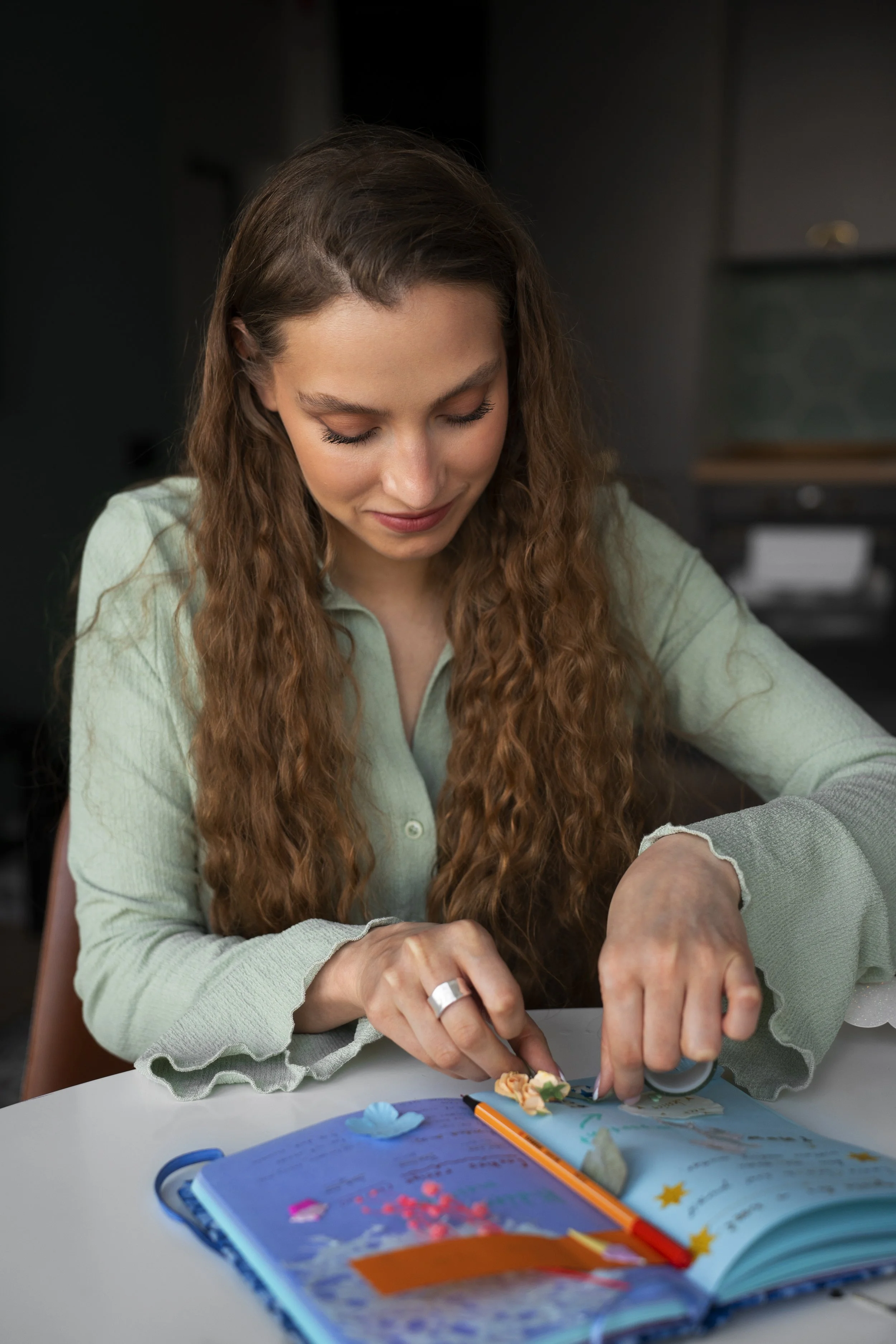 A woman with long curly hair sitting at a table, looking down at an open scrapbook with colorful decorations and stickers.