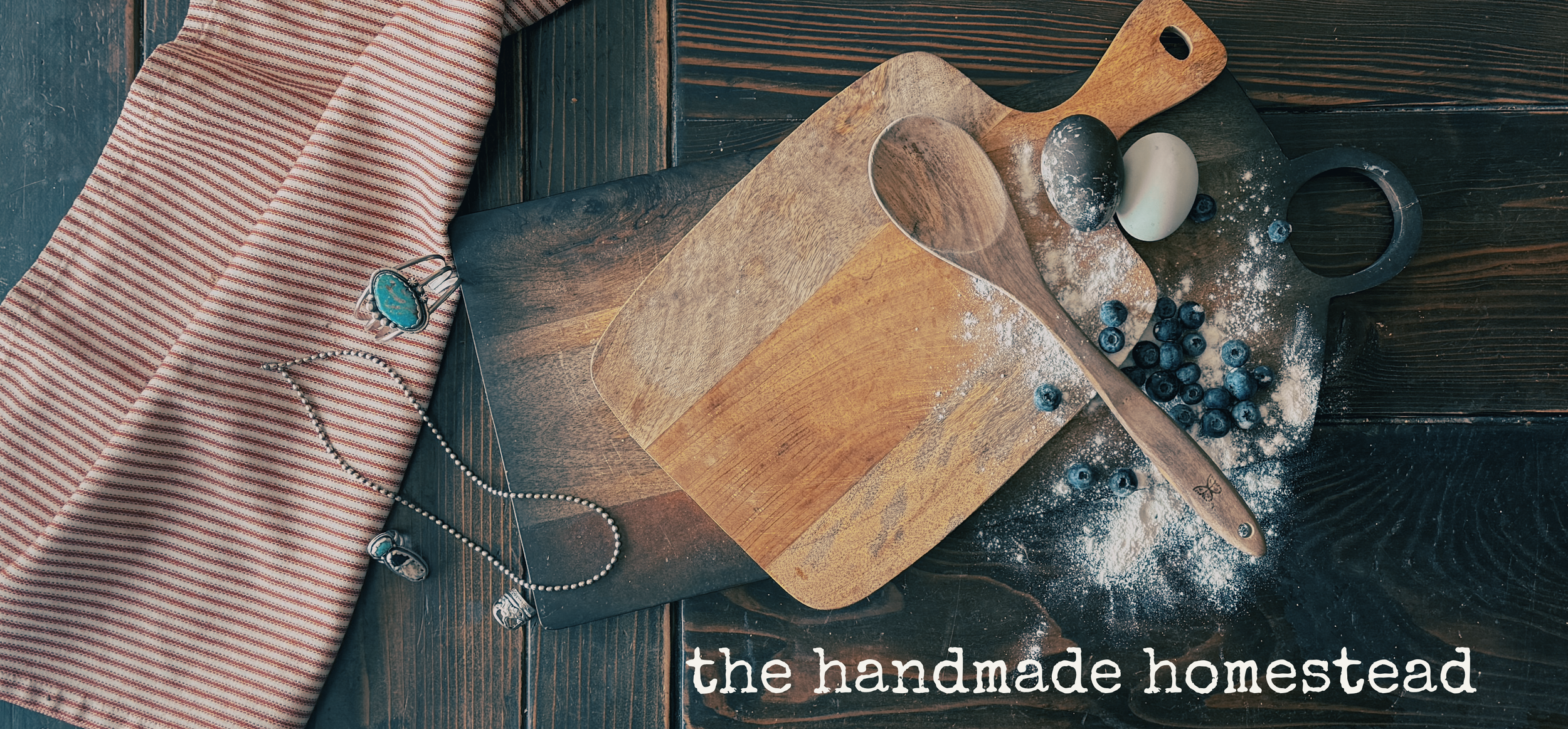 A rustic wooden table with a striped red and white cloth, a cutting board, a spoon, a black bowl, blueberries, eggs, and flour, with the text 'the handmade homestead' at the bottom right.