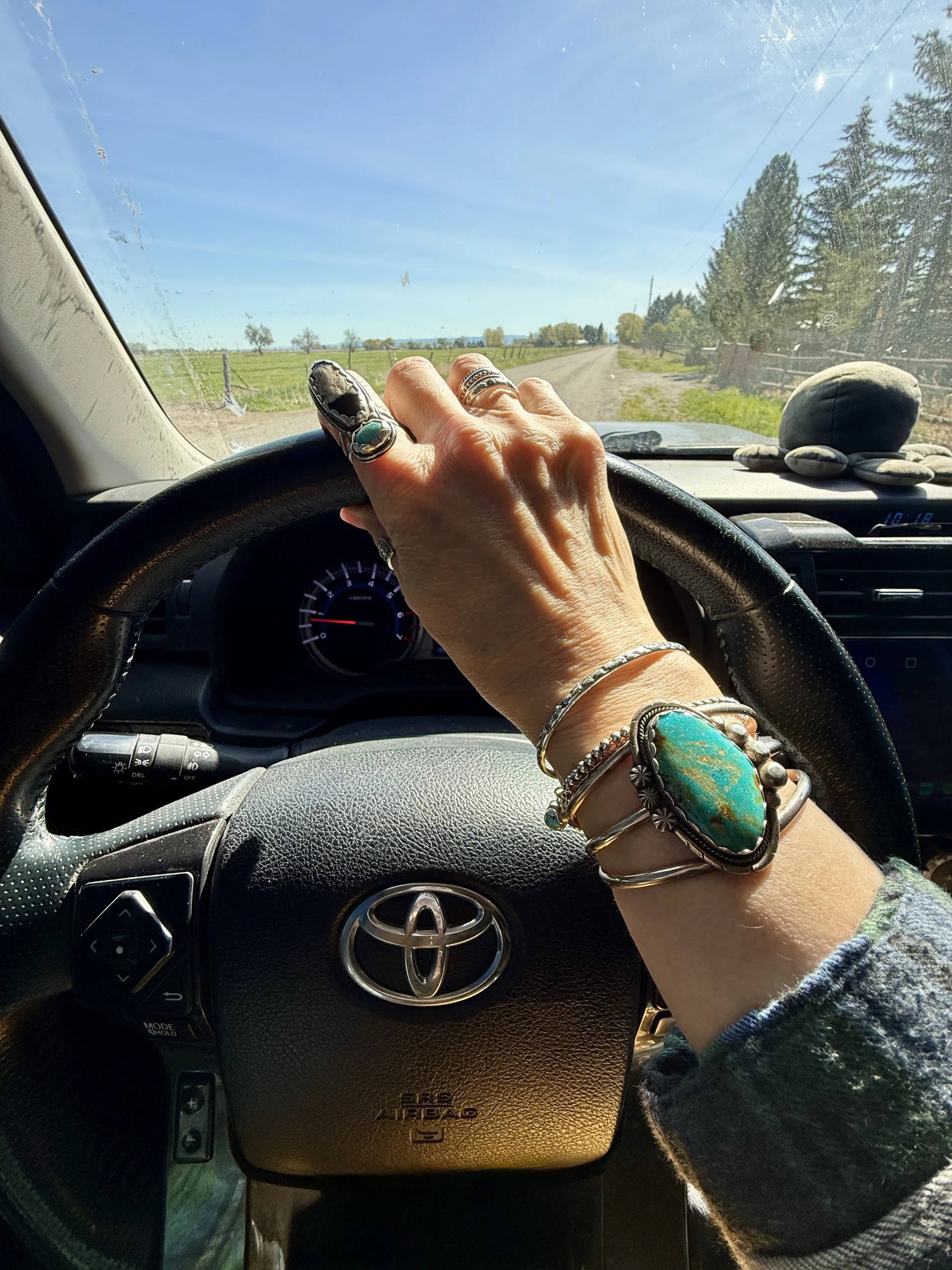 A close-up of a hand wearing handcrafted sterling silver and turquoise jewelry resting on a vehicle steering wheel, representing the M7 Auto Ranch and studio lifestyle.