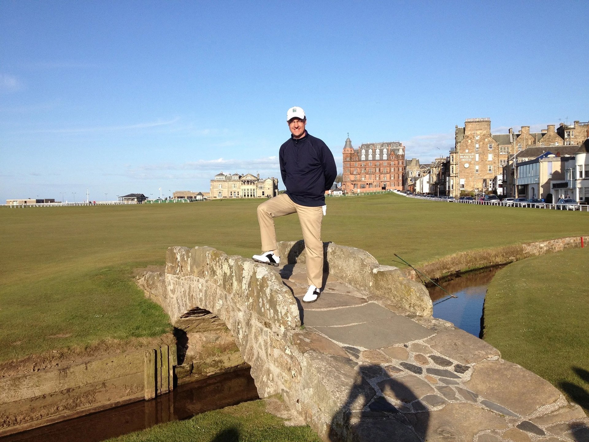 Man standing on a large rock on a golf course, with a row of historic buildings and a blue sky in the background.