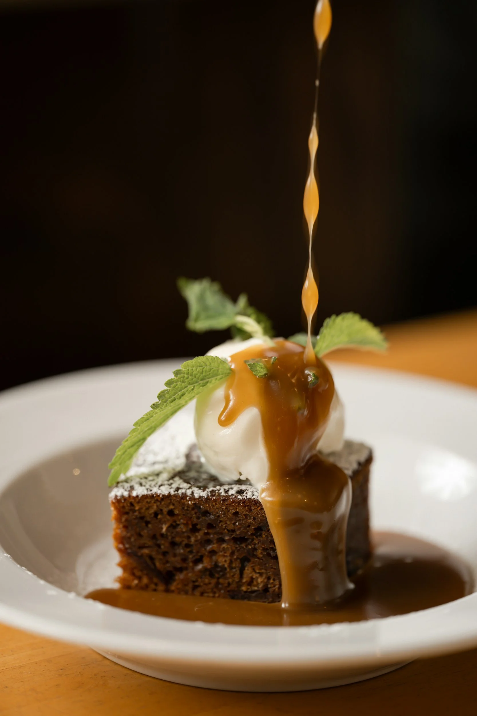 A slice of chocolate cake topped with a dollop of whipped cream, mint leaves, and caramel sauce, served on a white plate.