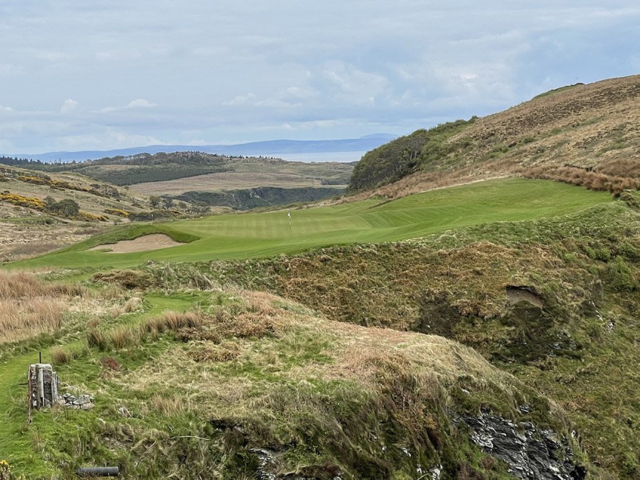 Scenic view of a golf course surrounded by rolling hills and lush green landscapes under a cloudy sky.