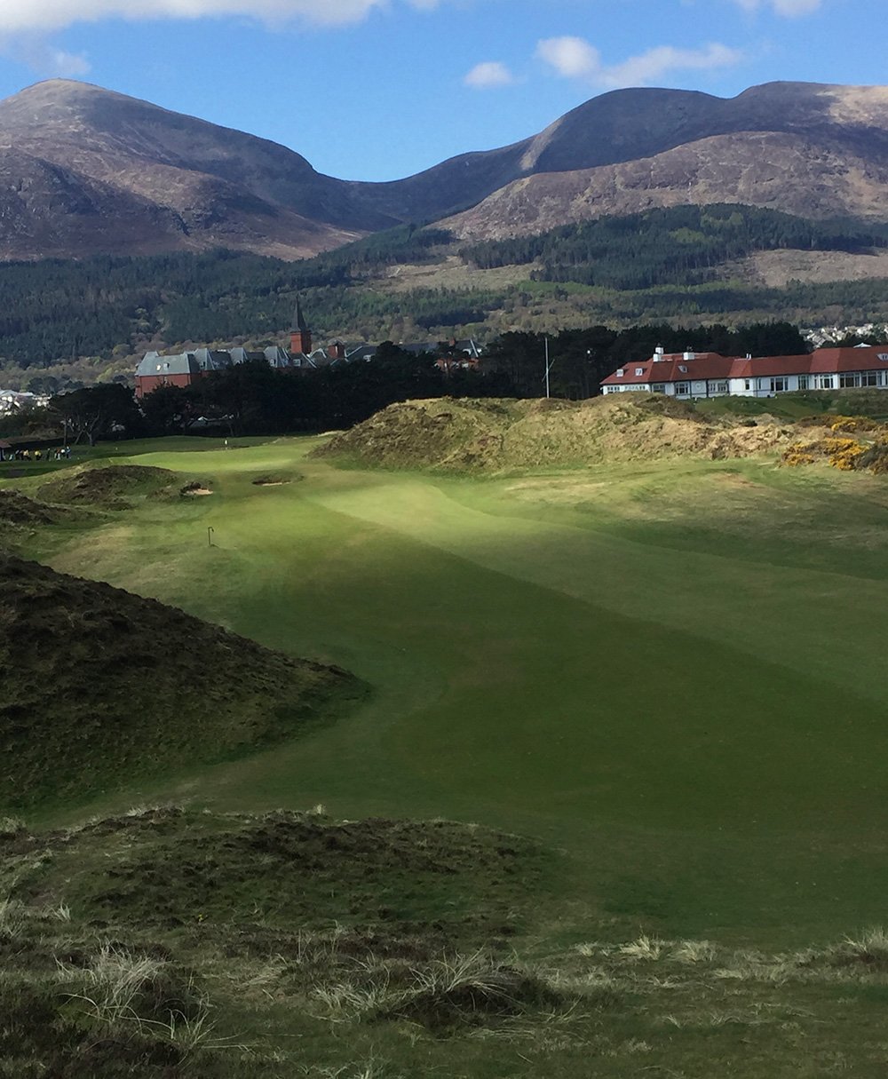 A golf course with green fairways and sand traps, surrounded by hills and mountains in the background, under a partly cloudy sky.