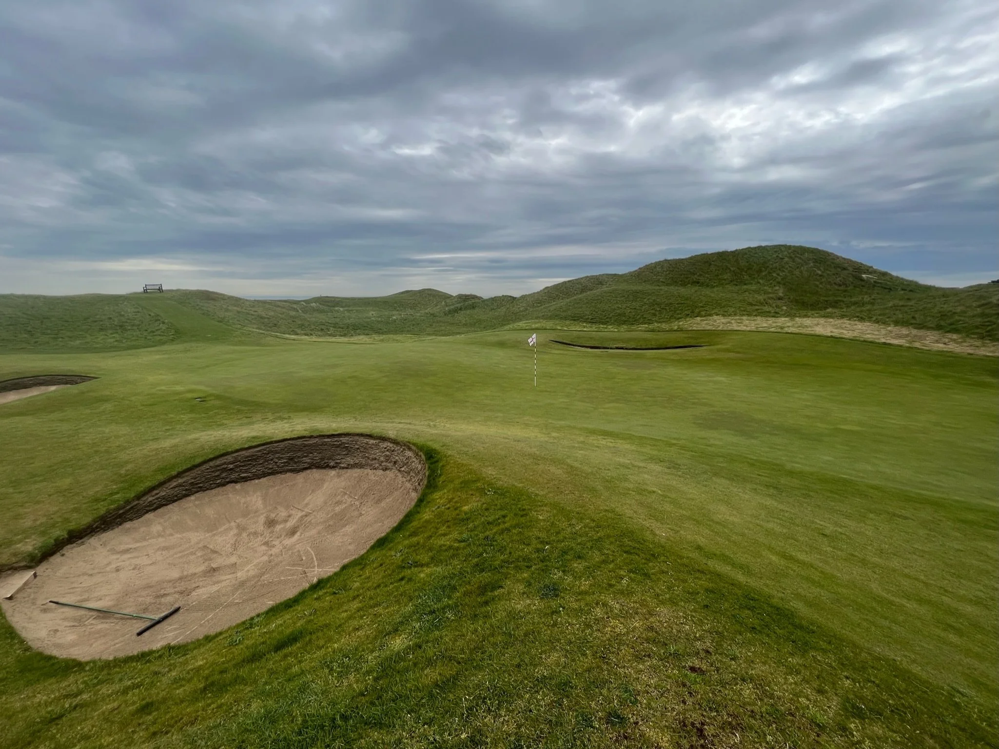 A golf course with a sand trap and a putting green under a cloudy sky, surrounded by green hills.