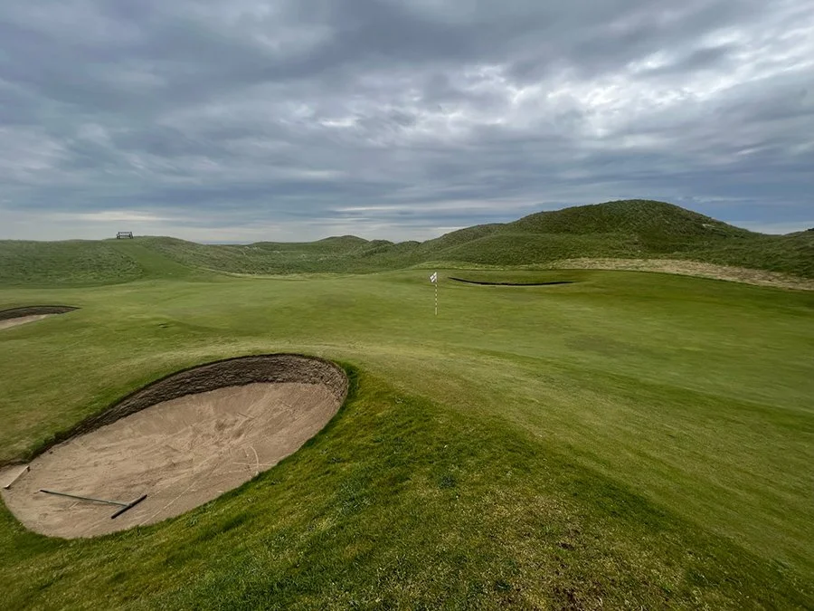 A golf course with a sand trap, green, flag, and rolling hills under a cloudy sky.