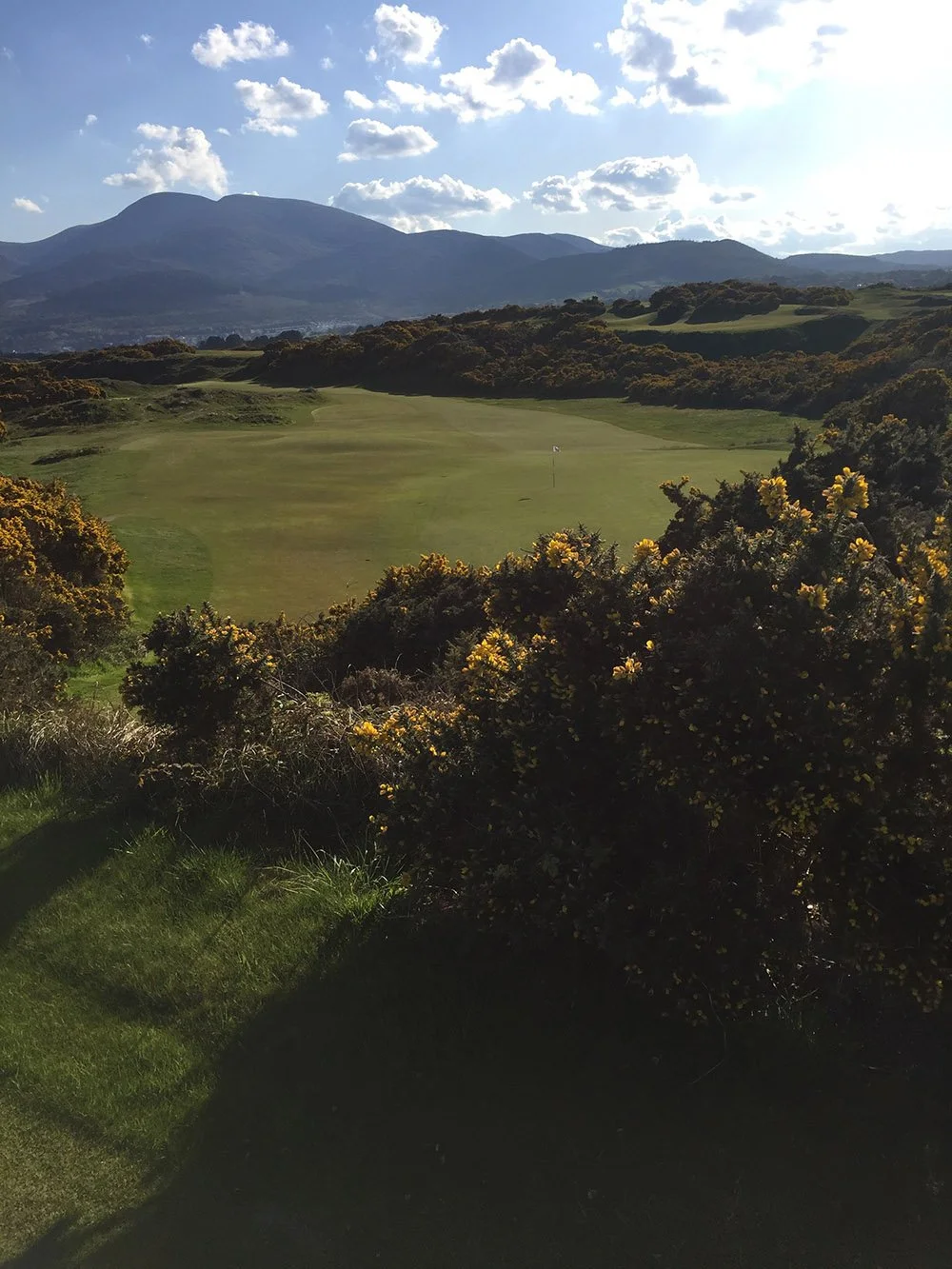 A golf course surrounded by bushes with mountains in the background under a partly cloudy sky.