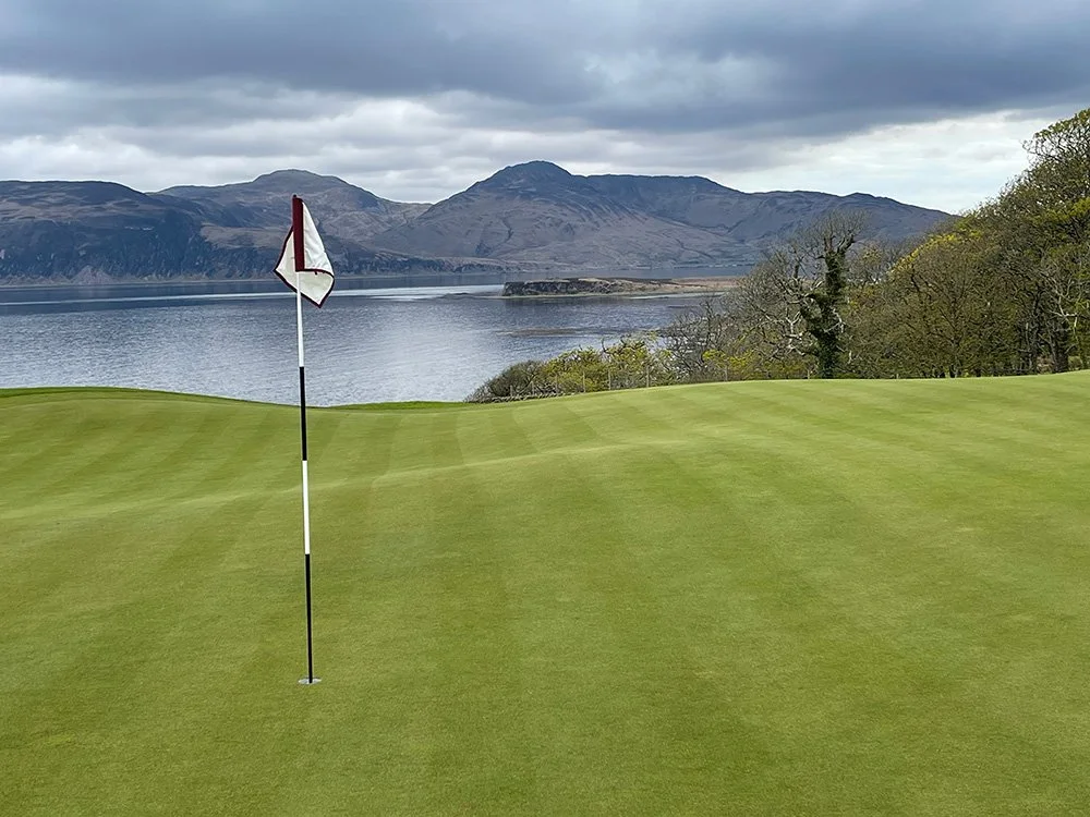 Golf course green with flagstick, overlooking a body of water and mountain range under cloudy sky.