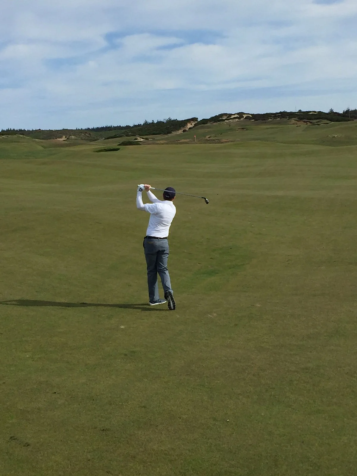 A person in a white shirt, gray pants, and a black cap is playing golf on a course with green grass and rolling hills in the background. The person is swinging a golf club.