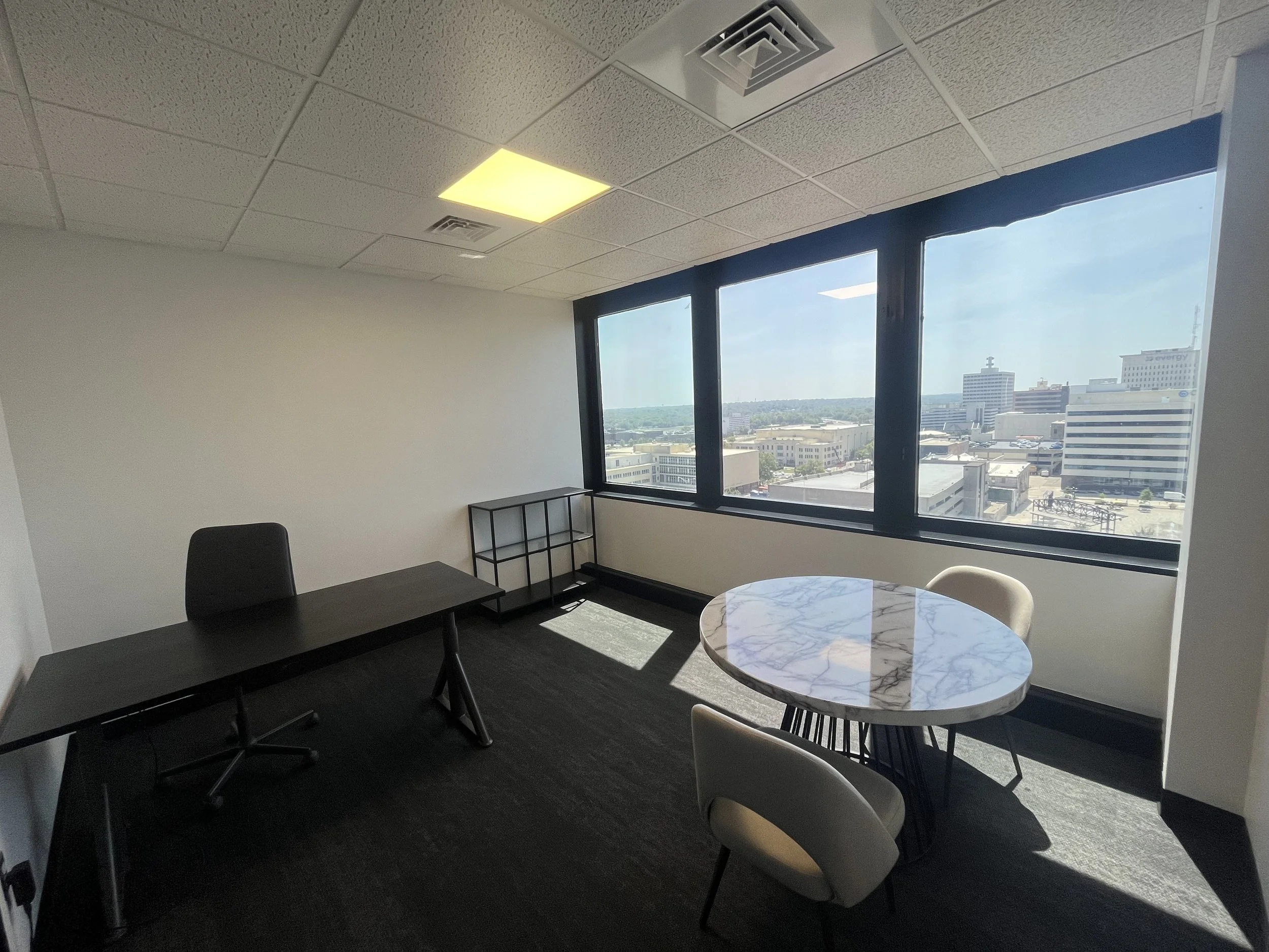 Empty office space with a black desk and chair, a small shelving unit, a round marble table with two chairs, large windows with cityscape view, and ceiling lights.