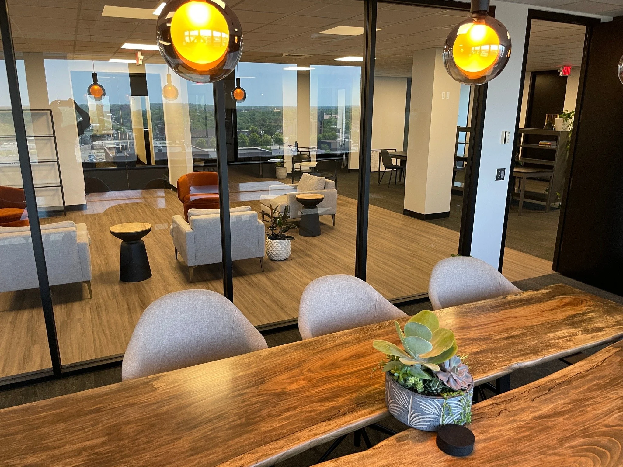 Modern office lounge with seating area and large windows showing city view, wooden flooring, hanging orange and yellow pendant lights, and an indoor potted succulent plant on a wooden table.
