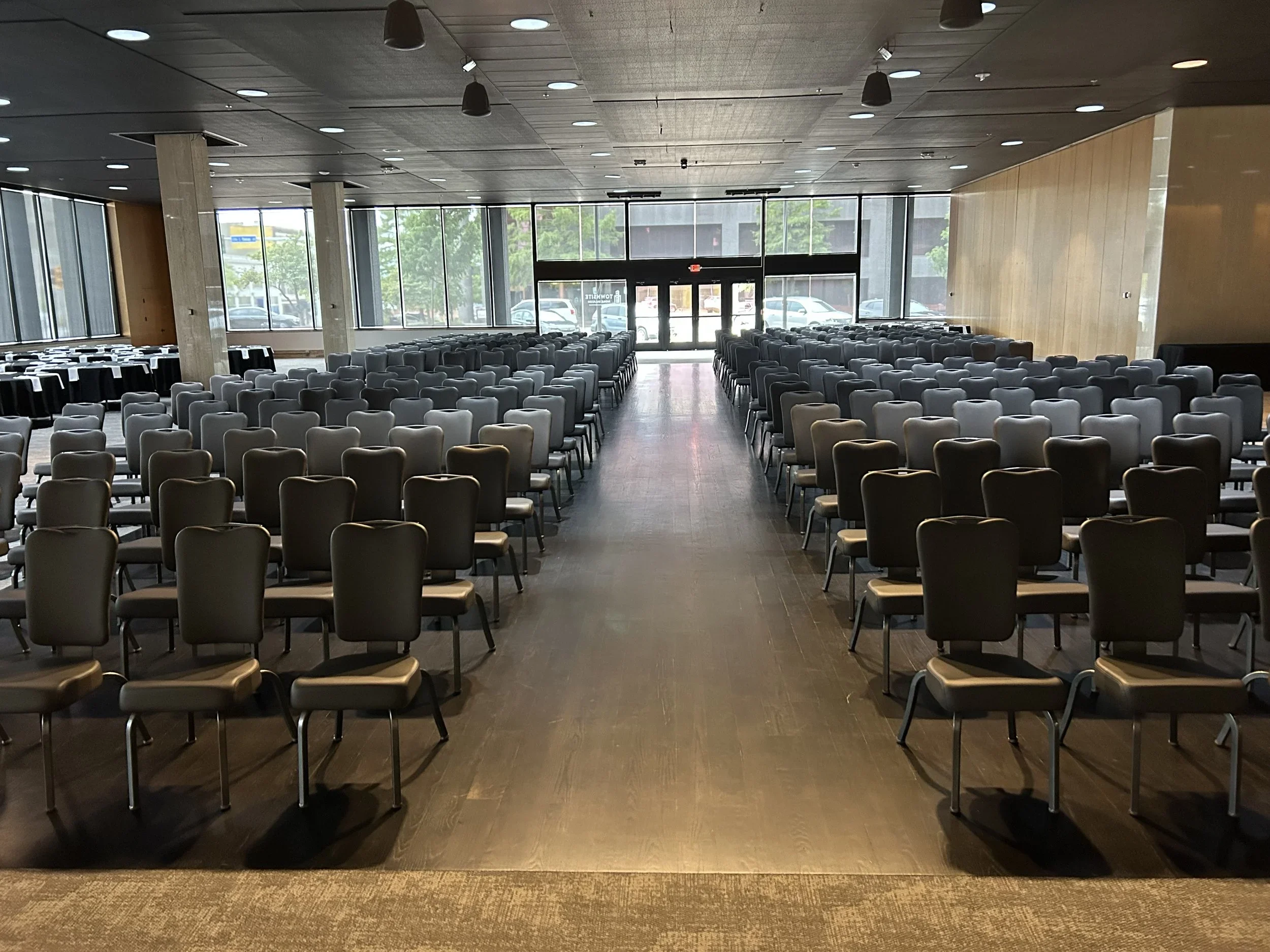 Empty conference room with rows of chairs facing towards the back entrance, large windows on the sides, and a wooden wall on the right side.