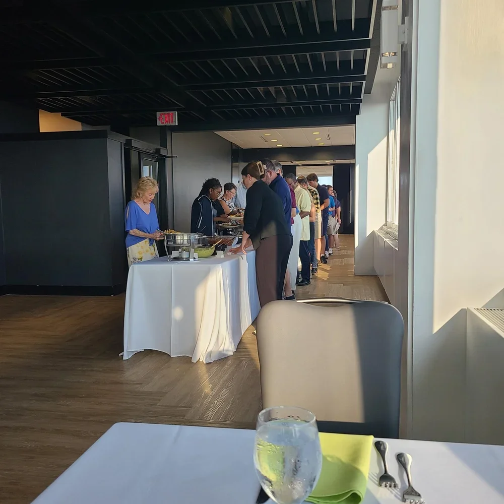 People lined up at a buffet table in a modern dining area, with sunlight streaming in through large windows, and a table in the foreground with a glass of water, a green napkin, and silverware.