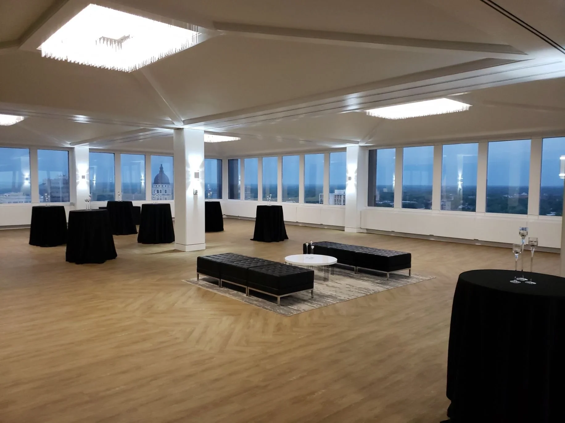 An empty event room with large windows, black cocktail tables with black tablecloths, black benches, and small white tables with glasses, set up for a gathering with a city view outside.