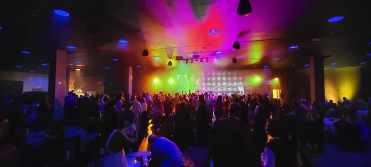 A crowded indoor concert with colorful stage lighting and people dancing.