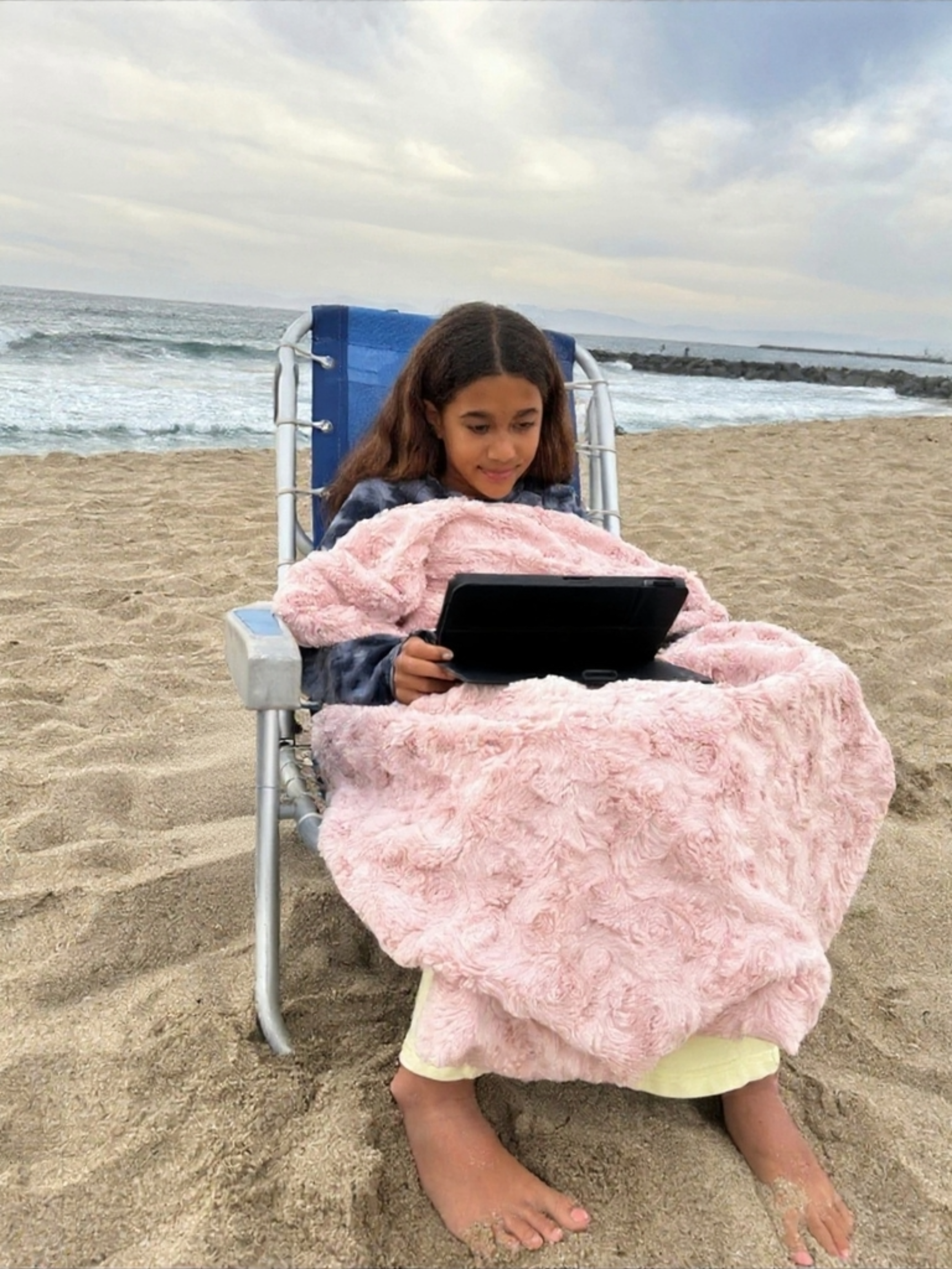 A girl sitting on a beach chair at the beach, looking at a tablet, covered with a pink blanket, with her legs and feet in the sand, at the shoreline with waves and a cloudy sky in the background.