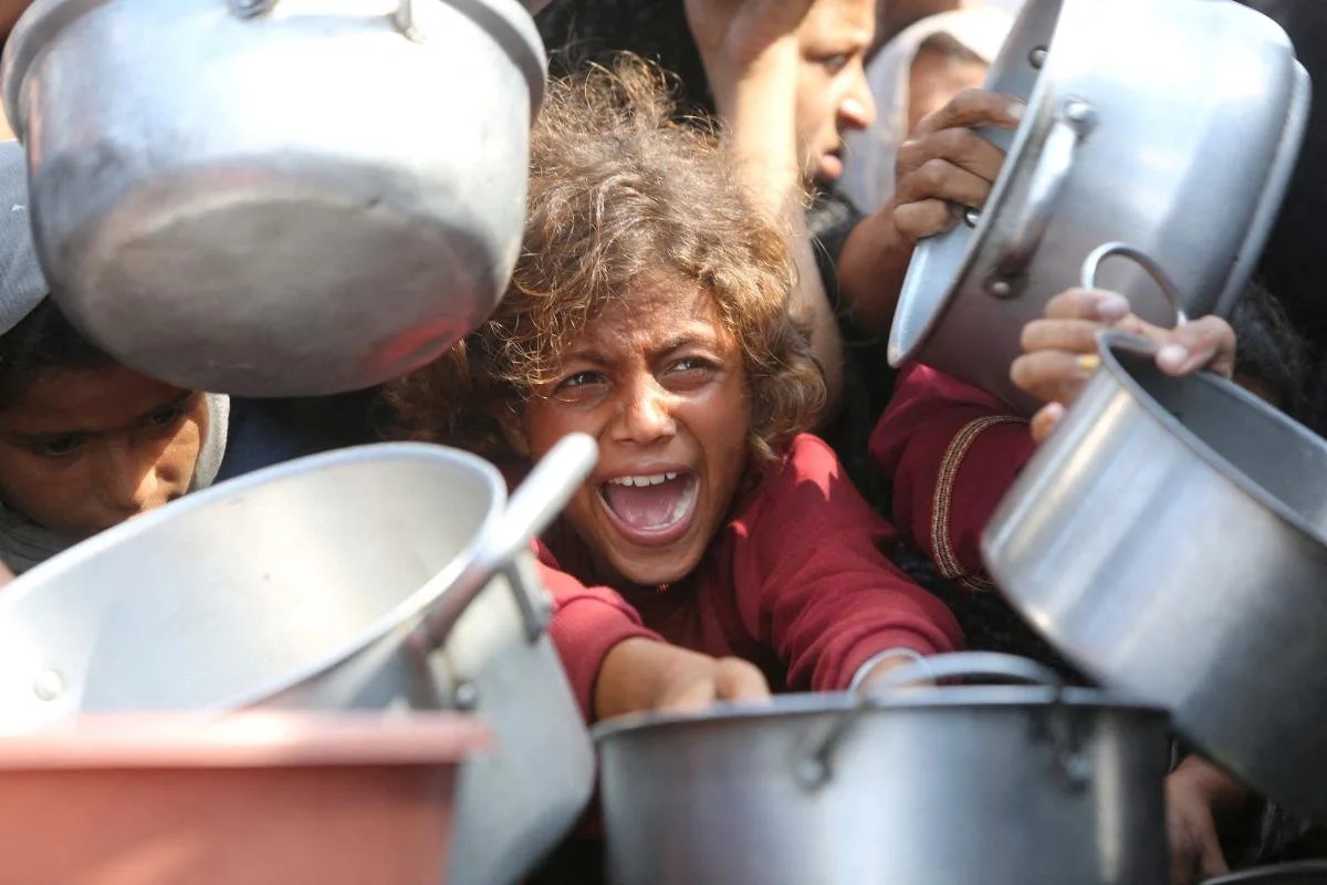 A young girl with curly hair and an expressive face, surrounded by metal pots and people, appears to be shouting or crying.