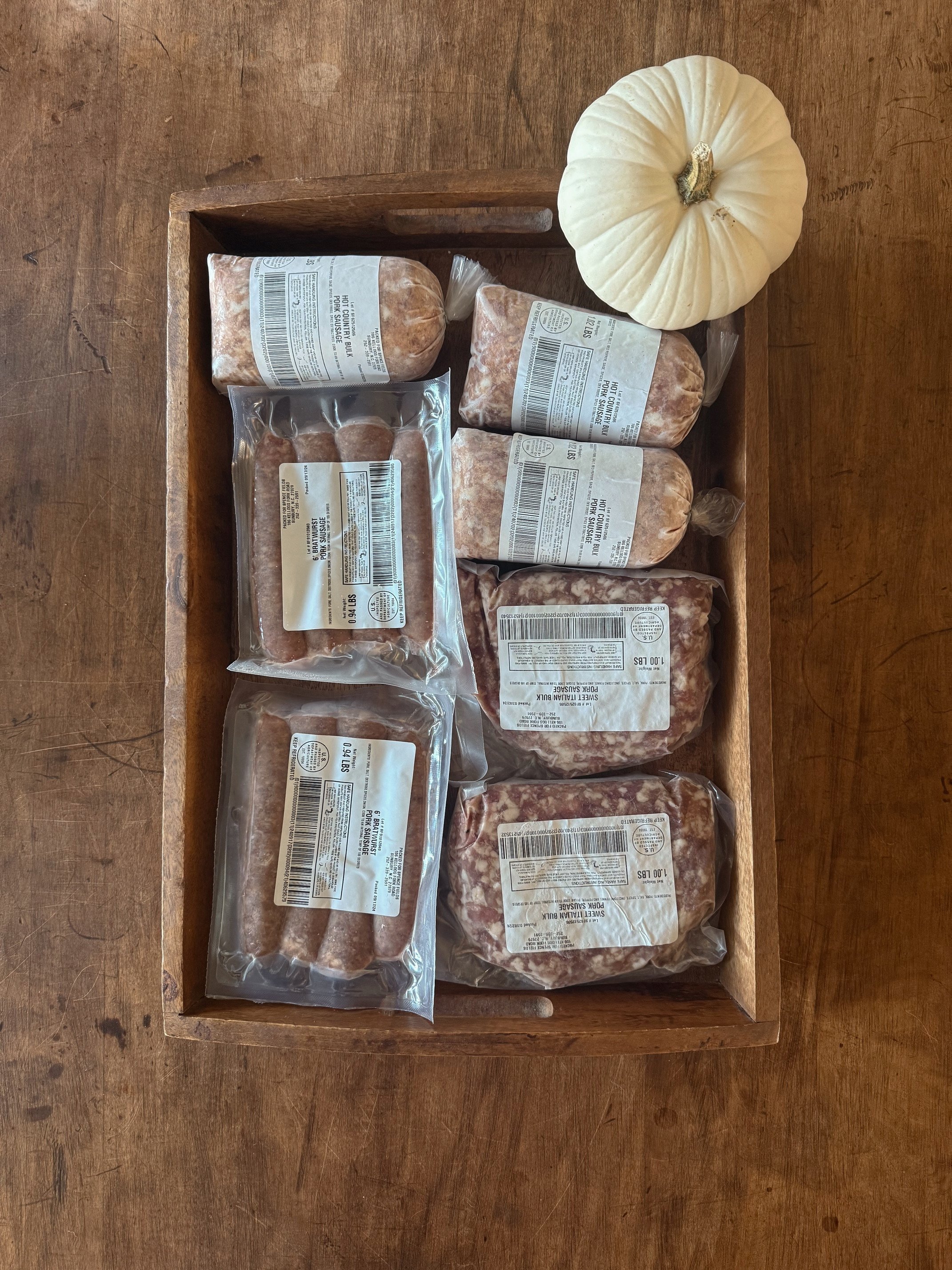 A wooden tray holding several packages of pork sausage and a white pumpkin on a wooden surface.