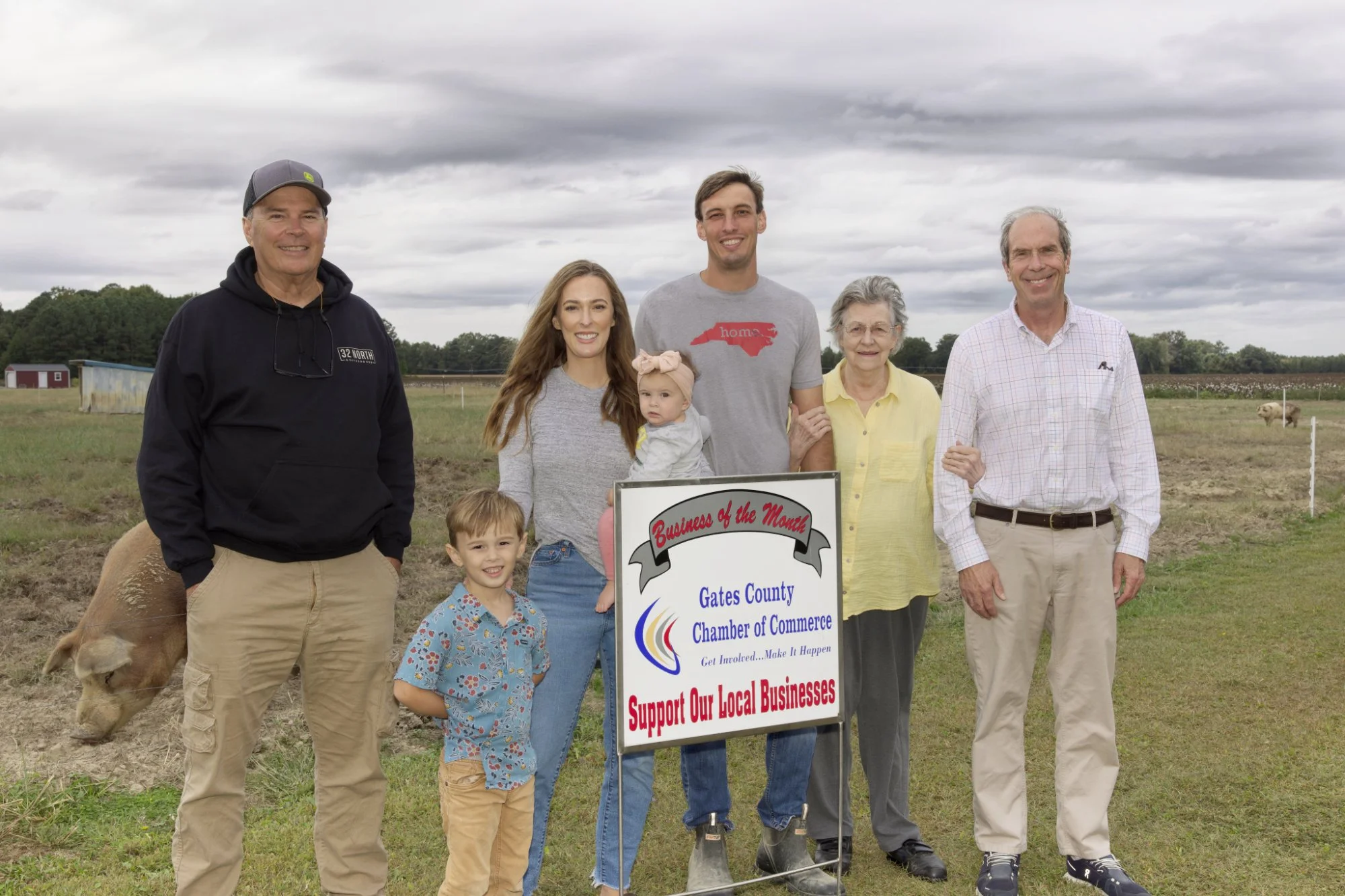 A diverse group of seven people, including adults and children, standing outdoors holding a sign for the Gates County Chamber of Commerce. The sign encourages supporting local businesses. The background shows a cloudy sky, farm buildings, and animals in a rural setting, indicating a community event or gathering.