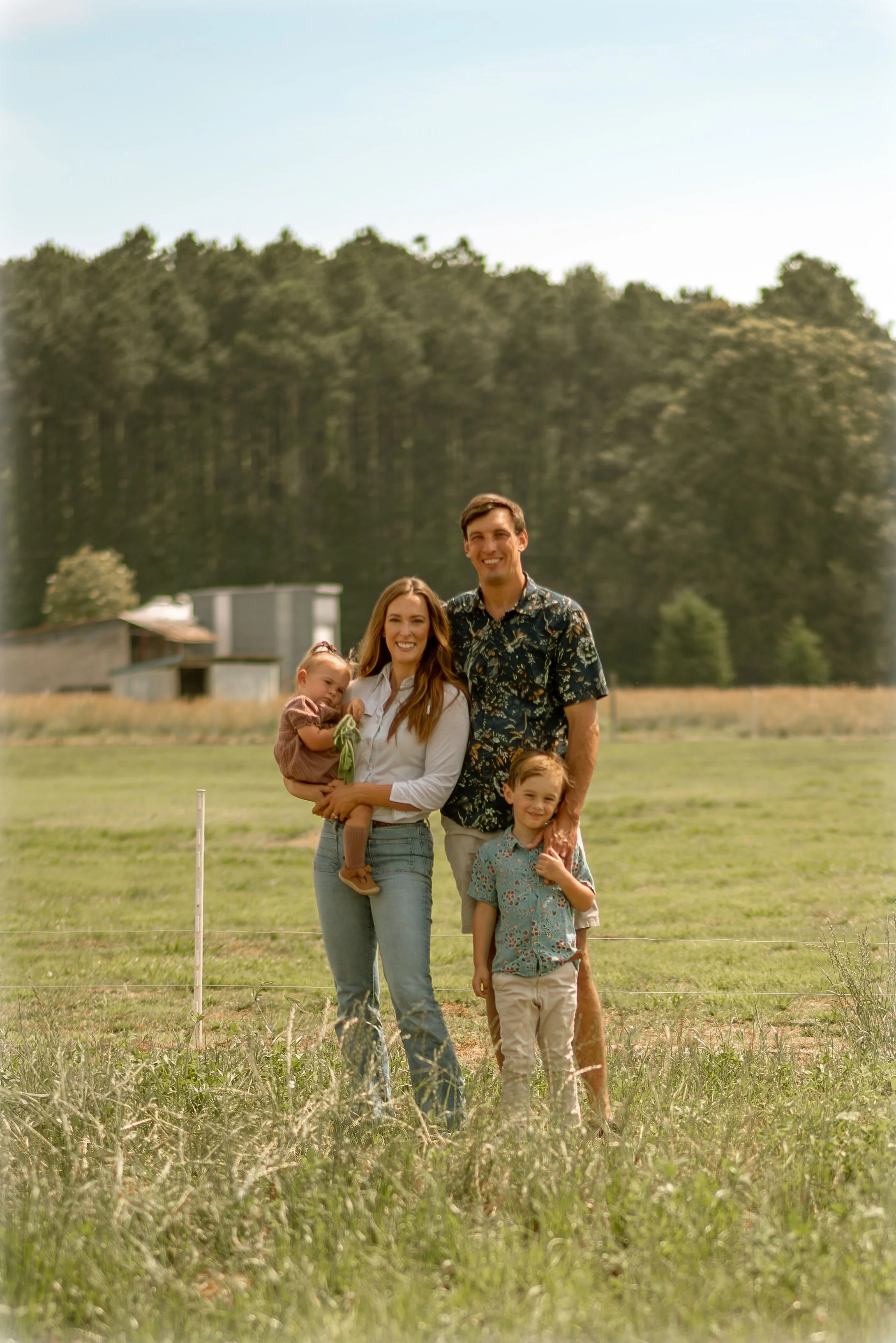 A family of five standing in a grassy field with trees in the background, smiling at the camera. The mother is holding a young girl, and two boys are standing in front with one of them at the father's side. The father is smiling and wearing a floral shirt.