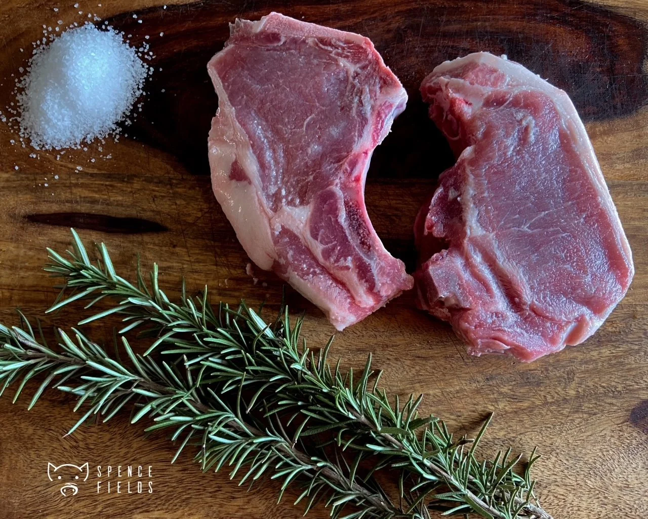 Two raw pork chops on a wooden cutting board with salt and a sprig of rosemary.