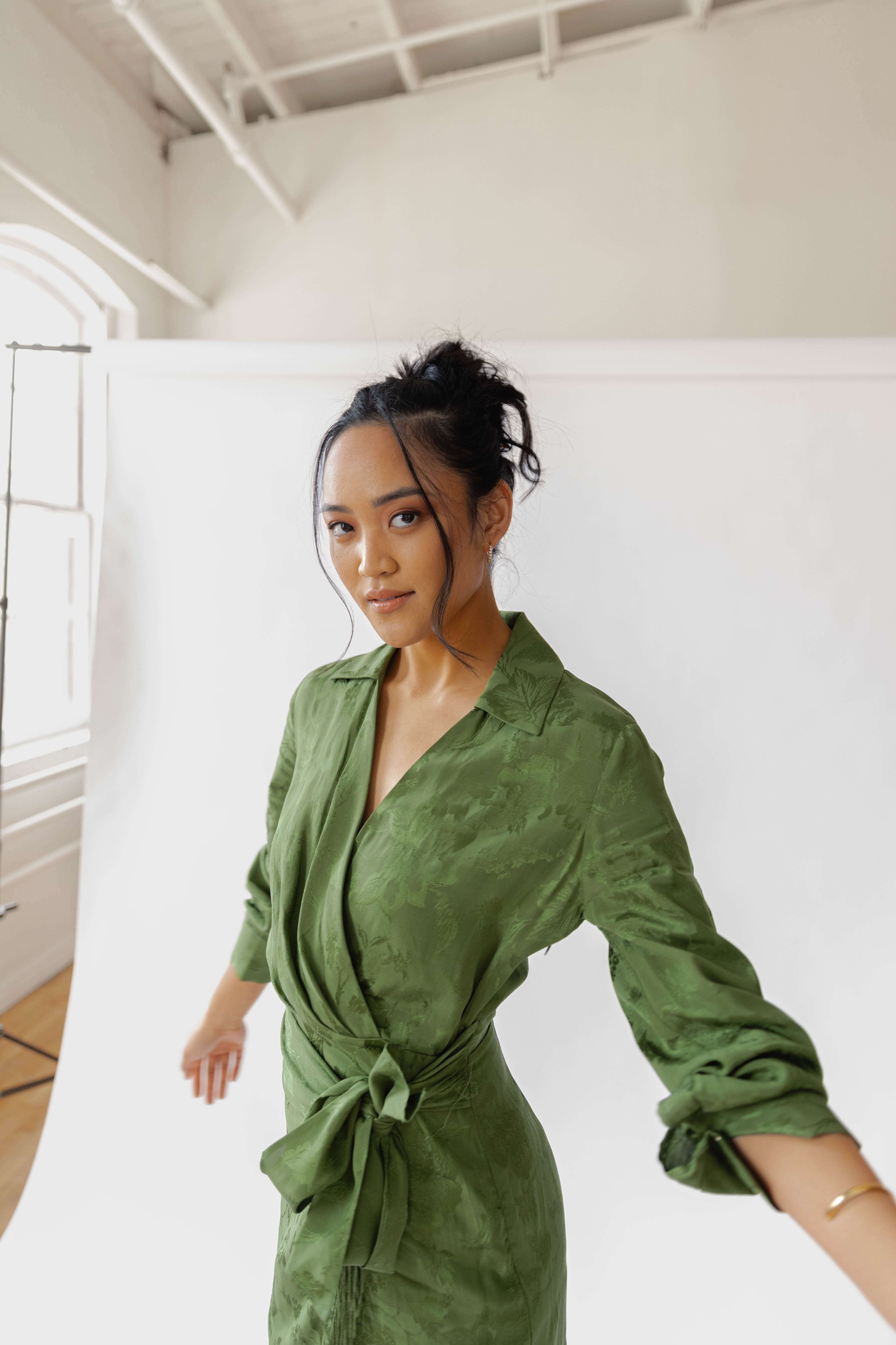 A woman wearing a green dress with a waist tie, posing in front of a white background in a photography studio.