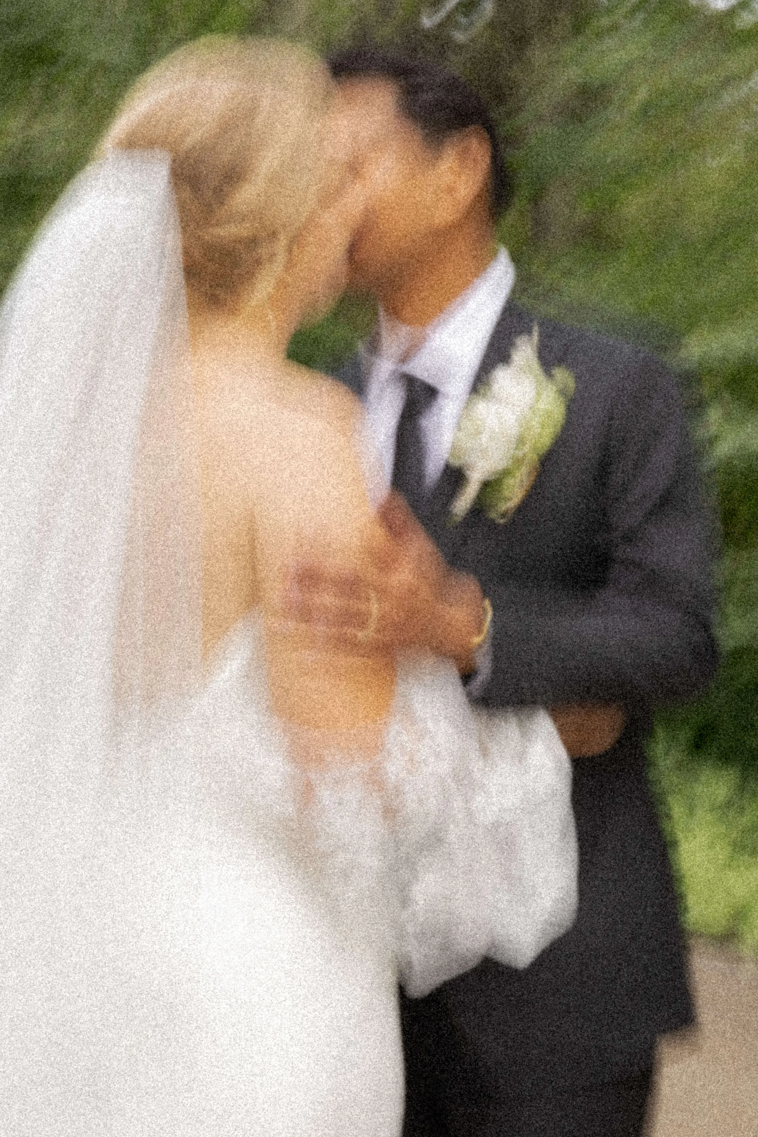 A newlywed couple kissing outdoors, with the bride in a white wedding dress and veil, and the groom in a dark suit with a boutonniere, surrounded by greenery.