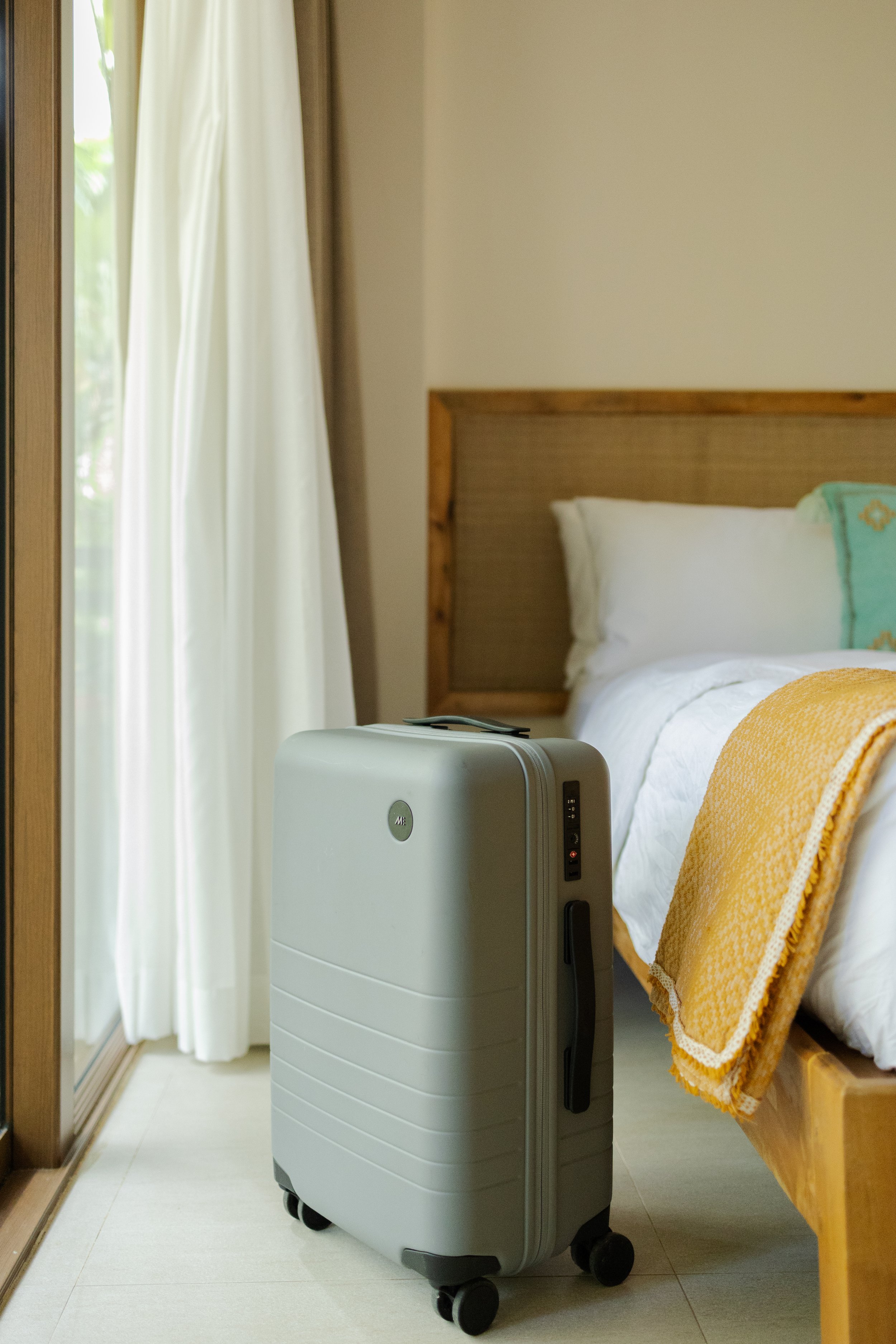 A bedroom with a suitcase on the floor next to the bed, a window with white curtains, a wooden headboard, and a yellow blanket on the bed.