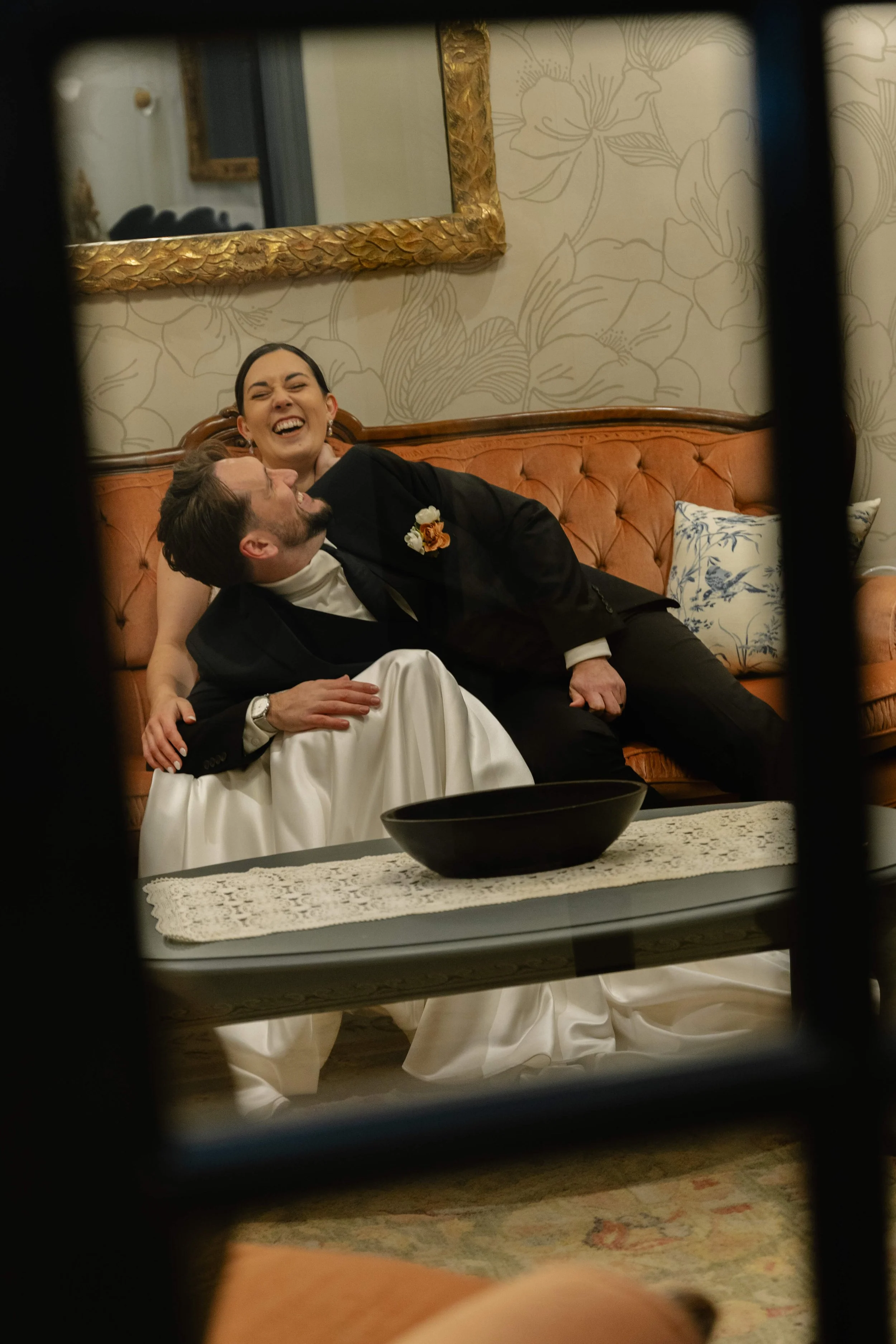 A couple sharing a joyful moment on a vintage style sofa, one in a wedding dress and the other in a black outfit, in a warmly decorated room.