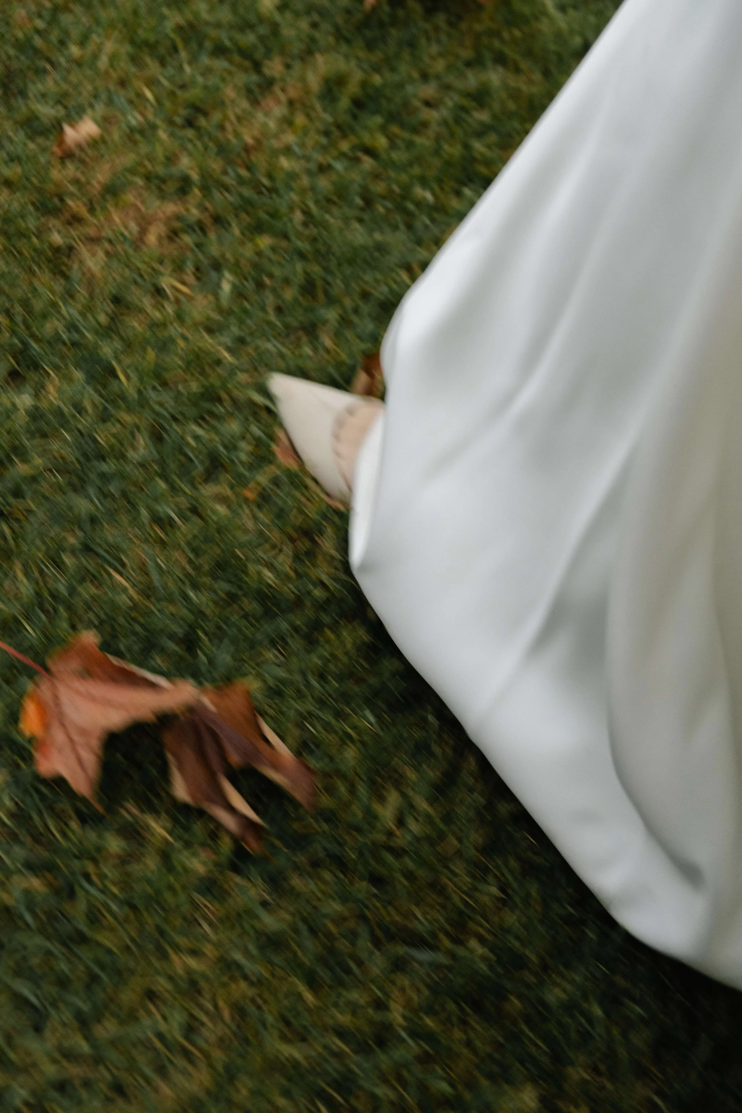 Close-up of a person's white clothing, possibly a dress or coat, walking on grass with fallen autumn leaves.