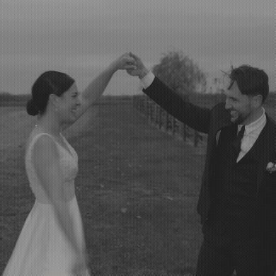A couple, dressed in wedding attire, dancing outdoors in a field with a fence and trees in the background, smiling and holding hands.