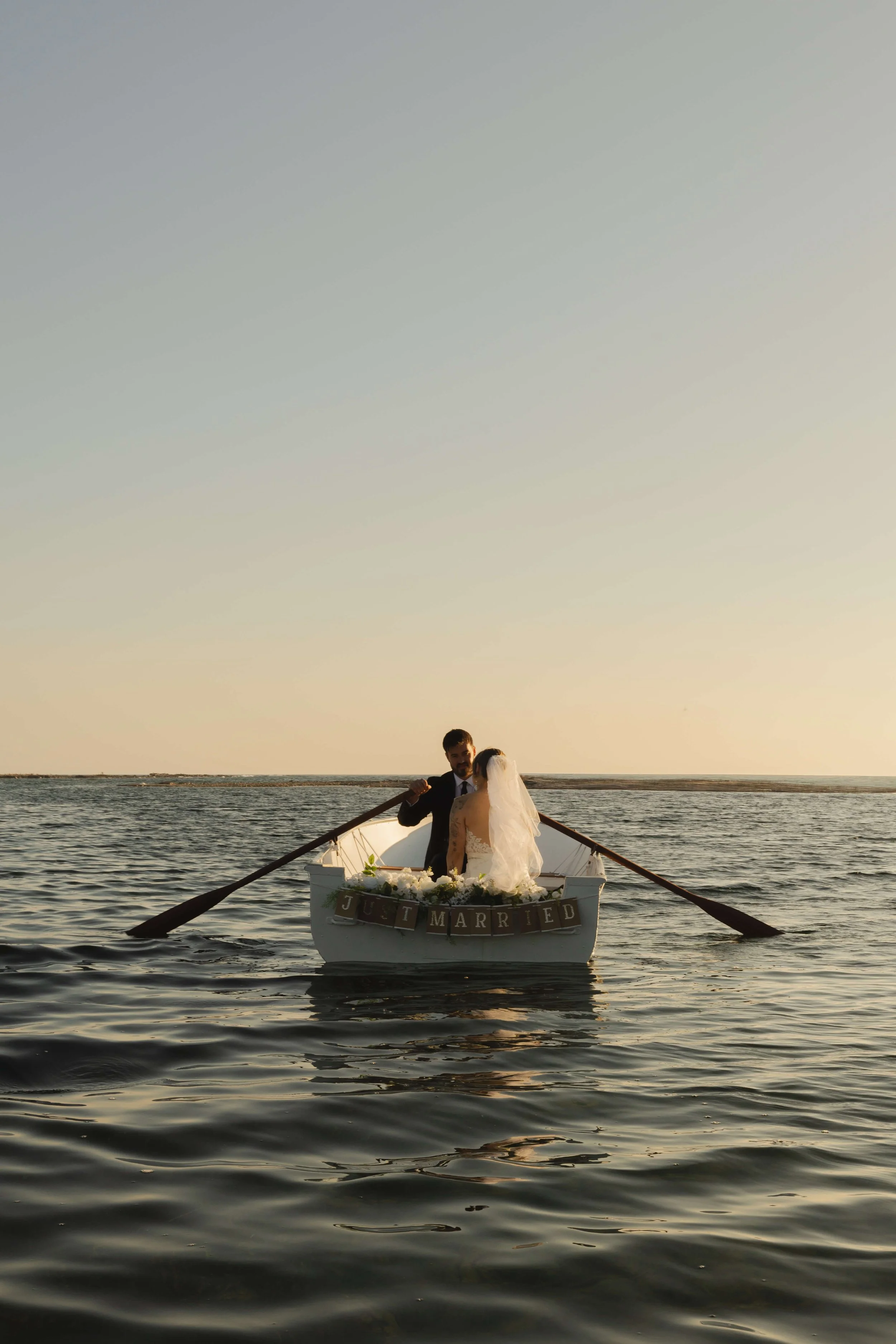 A newlywed couple sitting in a small boat on the water at sunset, with a "Just Married" sign on the back of the boat.