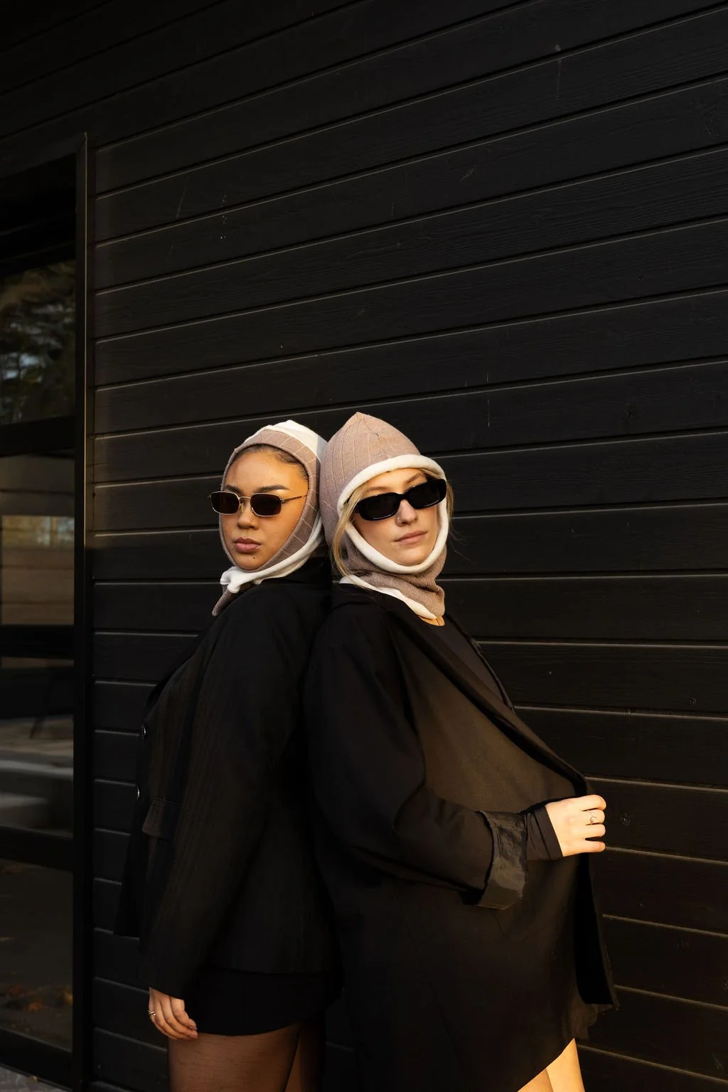 Two women wearing black jackets, beige hats, and sunglasses standing with their backs against a black wooden wall, facing forward.