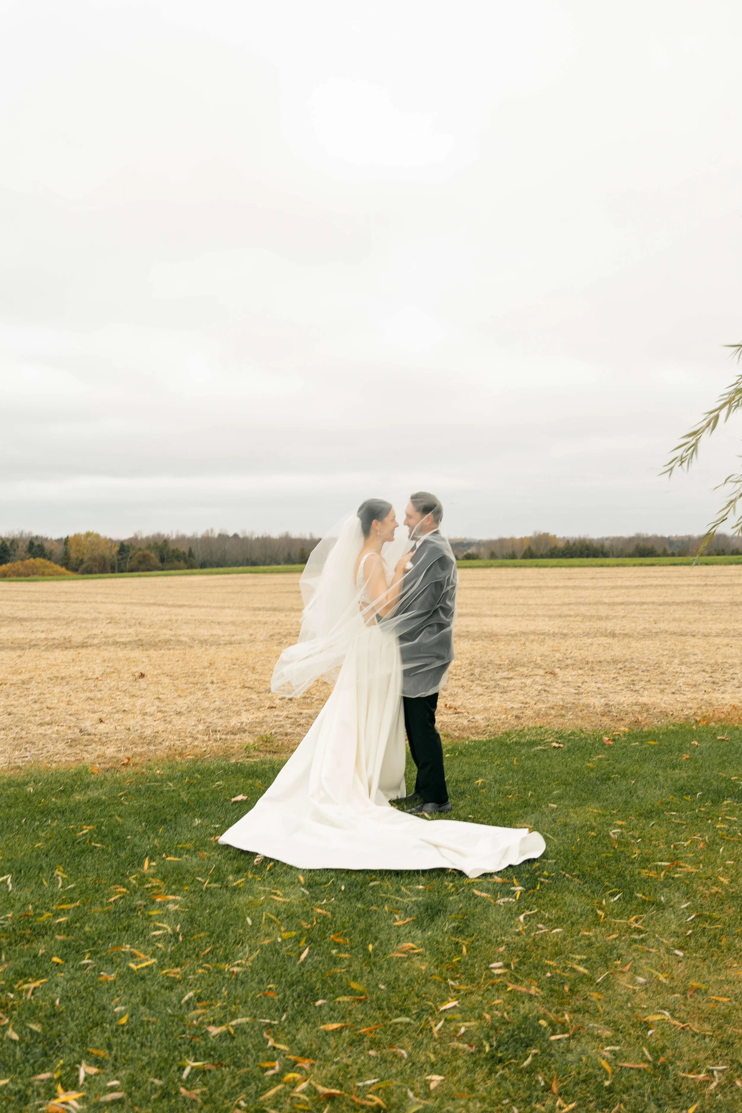 Bride and groom standing together outdoors on a cloudy day, facing each other with the bride wearing a white wedding dress and veil, the groom in a gray suit.