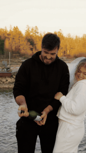 A man and a woman celebrating outdoors, opening a bottle of champagne near a waterfront with trees in the background.
