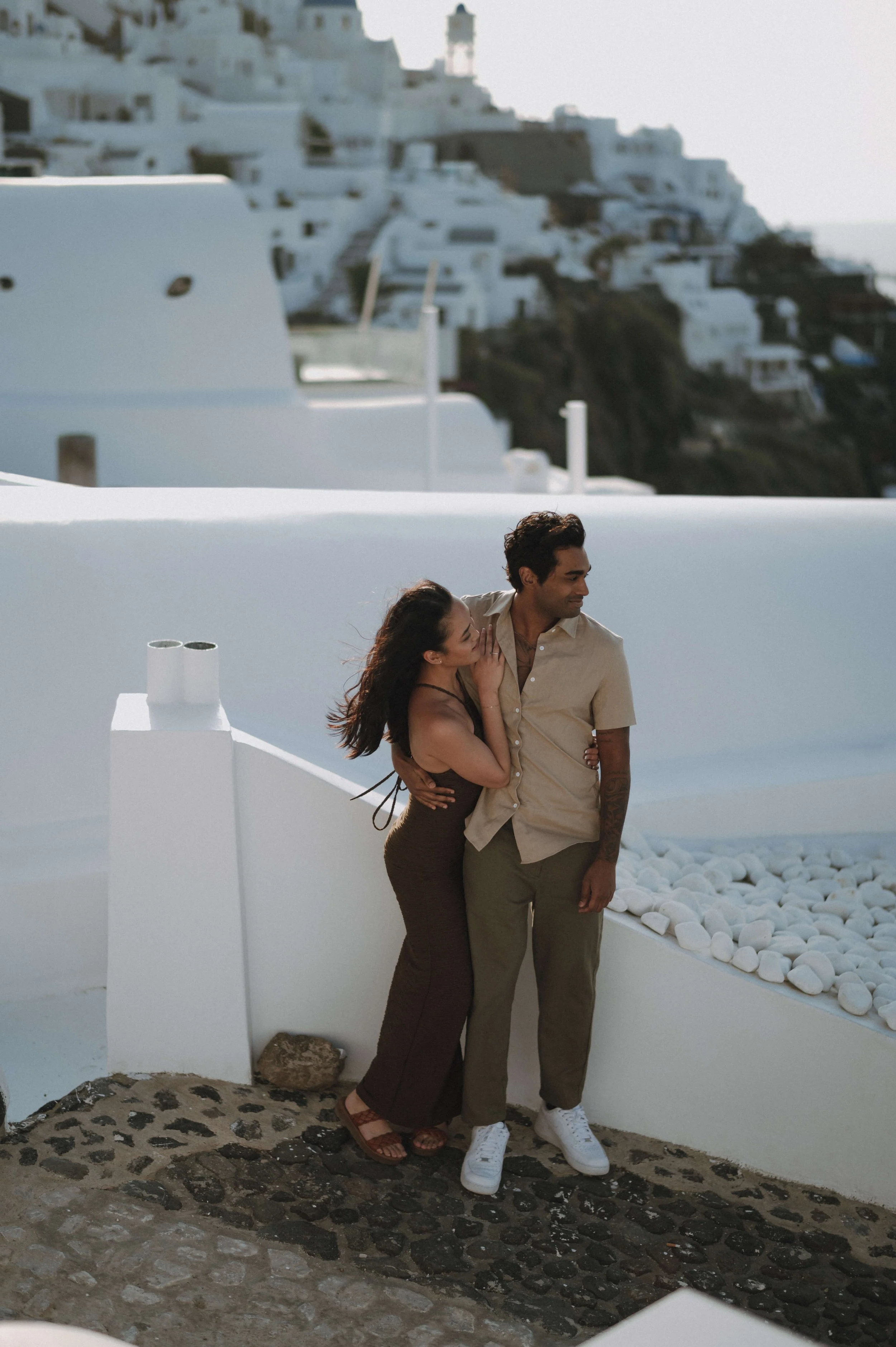 A couple stands close together in front of white buildings with white stones, with a hillside town backdrop, possibly Santorini, Greece.