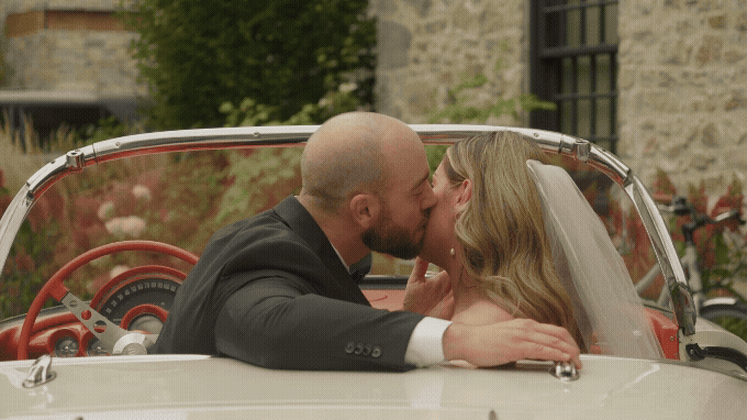 A newlywed couple sharing a kiss in a classic convertible car, with the groom in a suit and the bride in a wedding dress and veil, outdoors near a stone building and greenery.