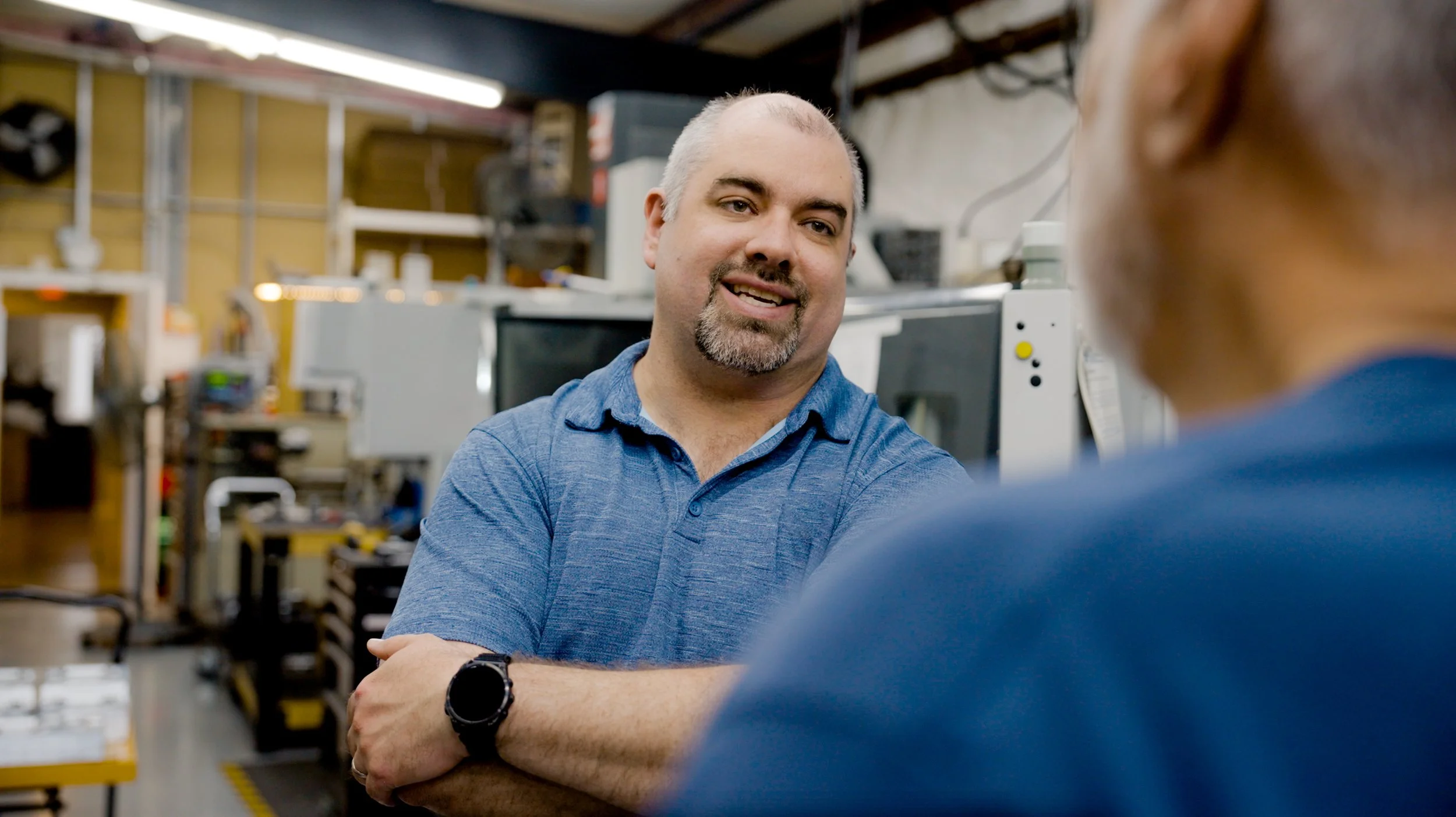Two men having a conversation in a workshop or factory, with one man smiling and wearing a blue shirt and smartwatch, the other partially visible from the back.