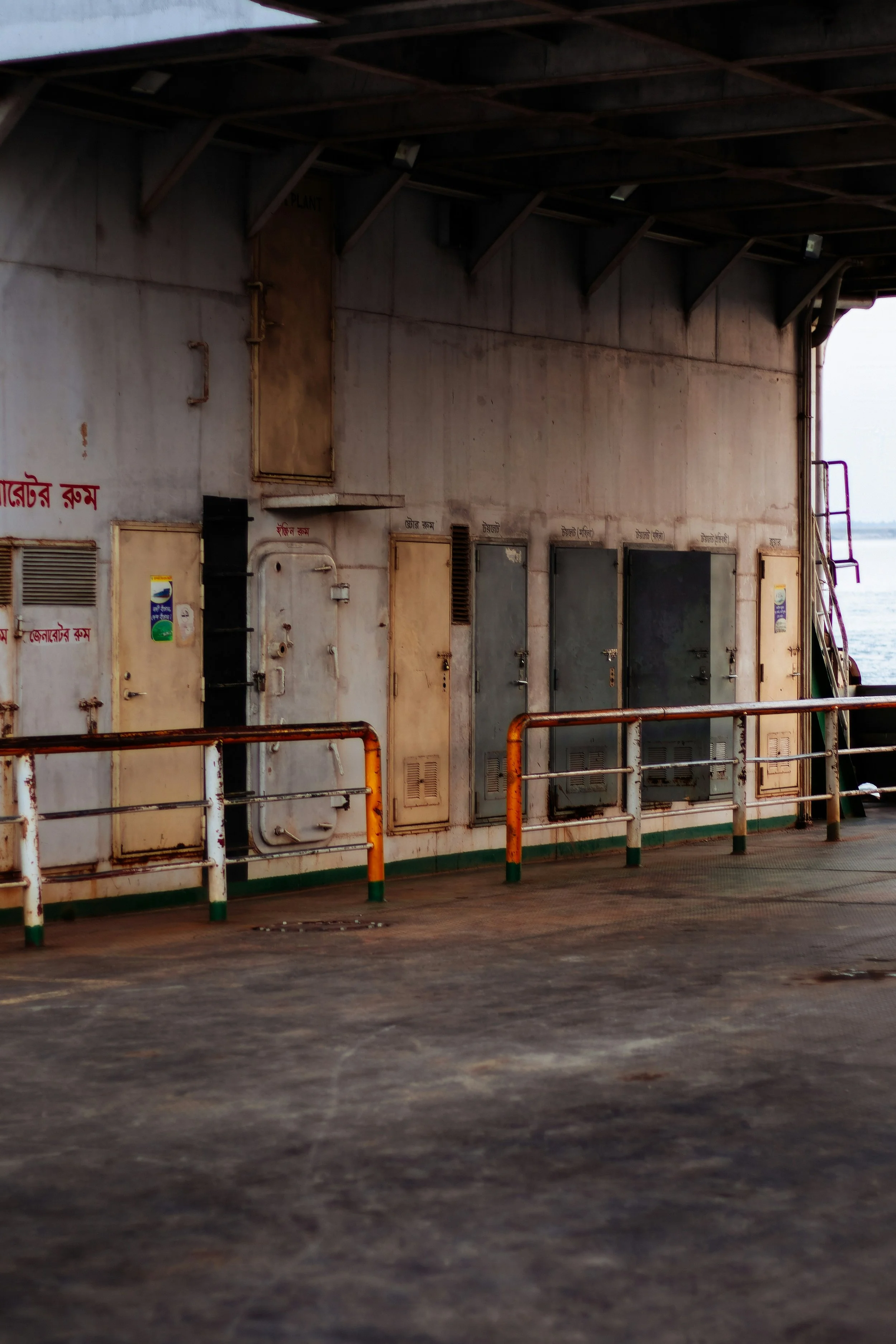 A section of the exterior of a boat or ferry, showing rusted metal doors and panels, with a railing in the foreground, and water visible in the background.