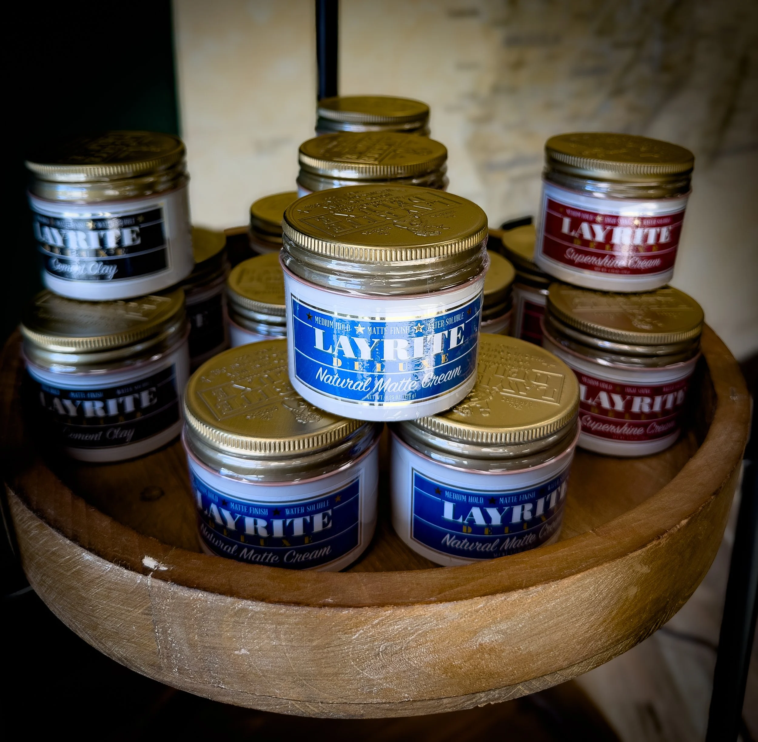 Several jars of Layrite Natural Matte Cream with gold lids, placed in a wooden bowl.