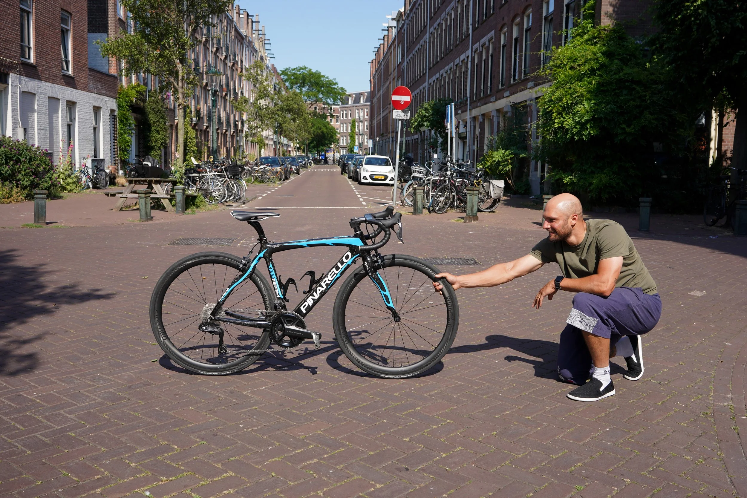 A man kneeling on one knee and holding a bicycle's front wheel on a brick street in a city with parked bicycles and cars, and residential buildings in the background.