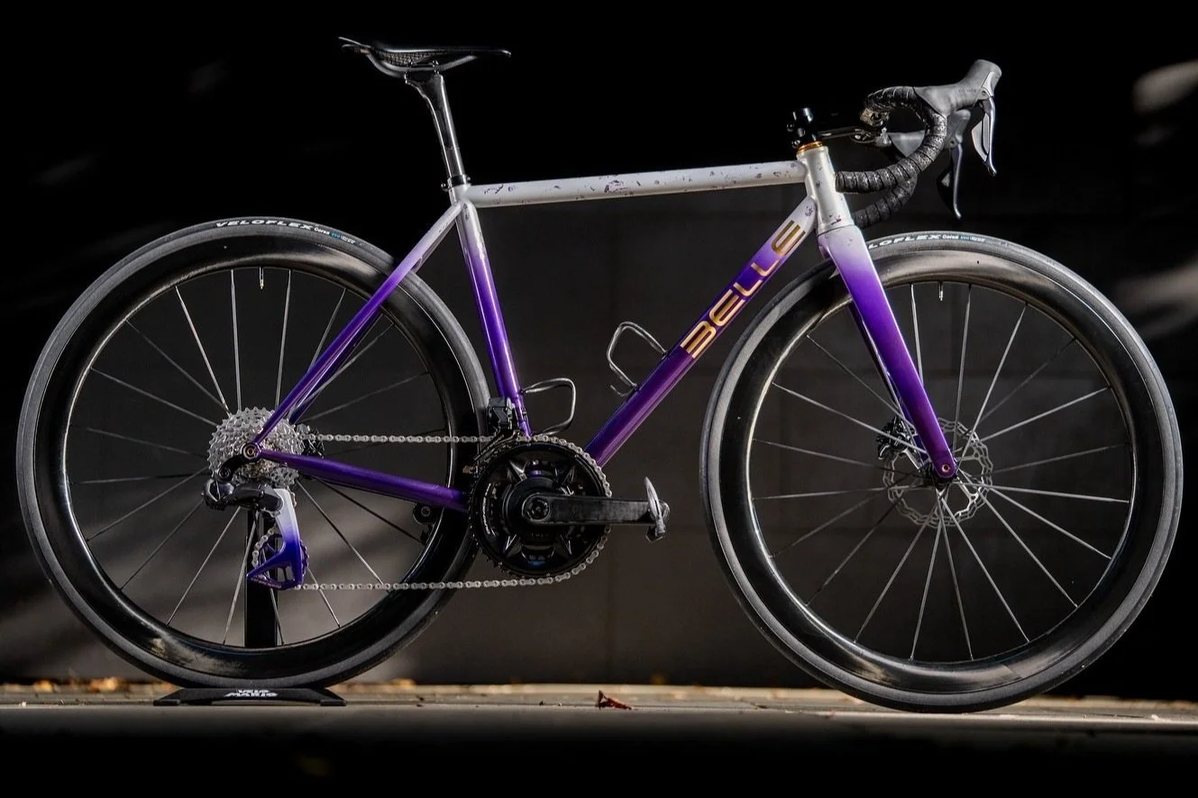 A purple and silver BMC road bicycle with drop handlebars and black tires, standing against a dark background.